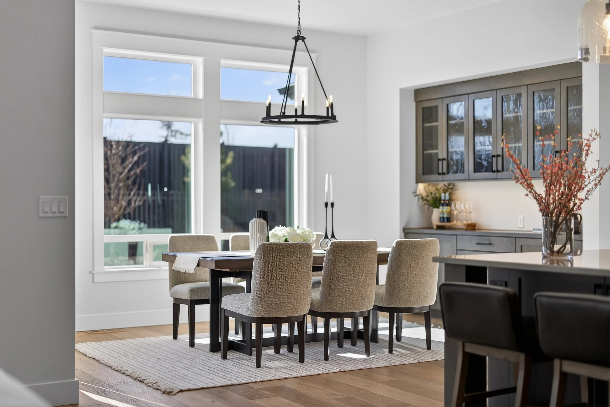 Dining area with a wooden table and beige upholstered chairs in a bright room with large windows, pendant light, and decorative items on the sideboard.