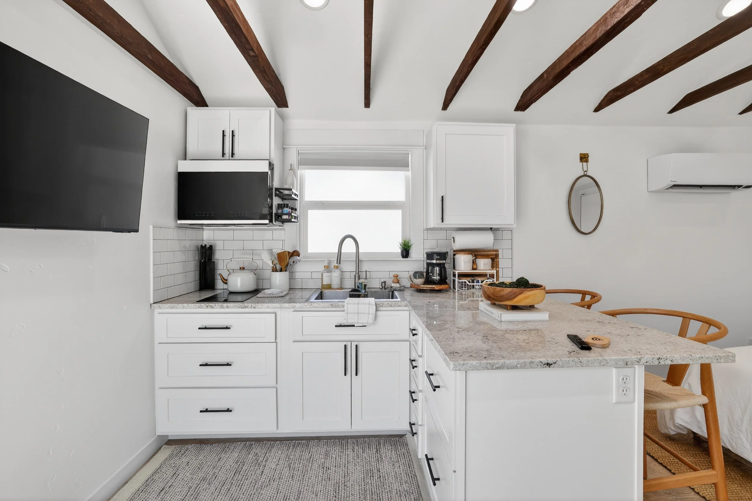 Modern kitchen with white cabinets, marble countertop, and wooden ceiling beams. Contains a window above the sink, a wall-mounted TV, and a small dining area with wooden chairs.