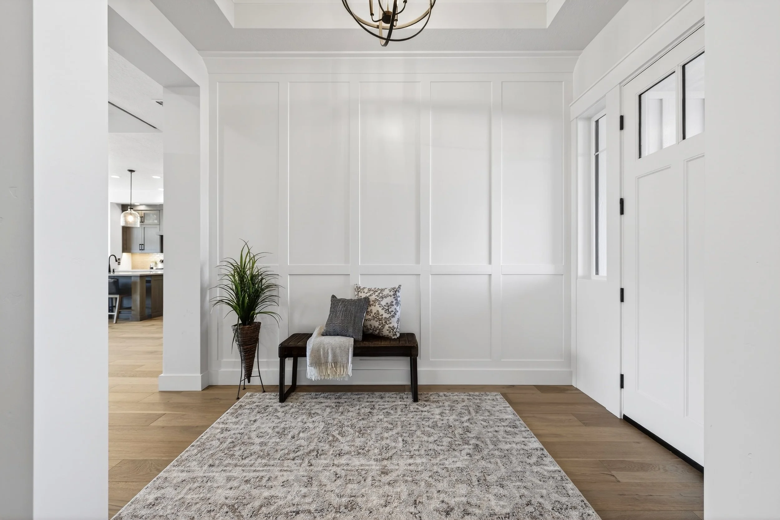 Modern white entryway with wooden bench, decorative pillows, indoor plant, rug, and a front door with glass panels.
