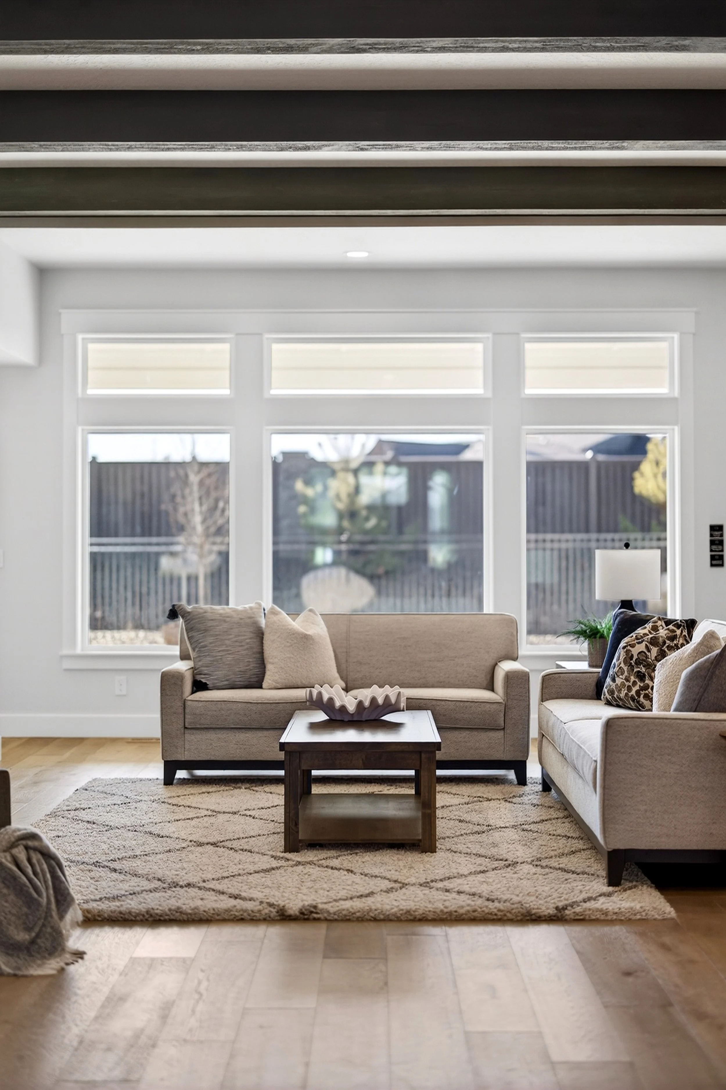 Living room with beige sofas, a wooden coffee table, large windows, a rug, and a lamp.