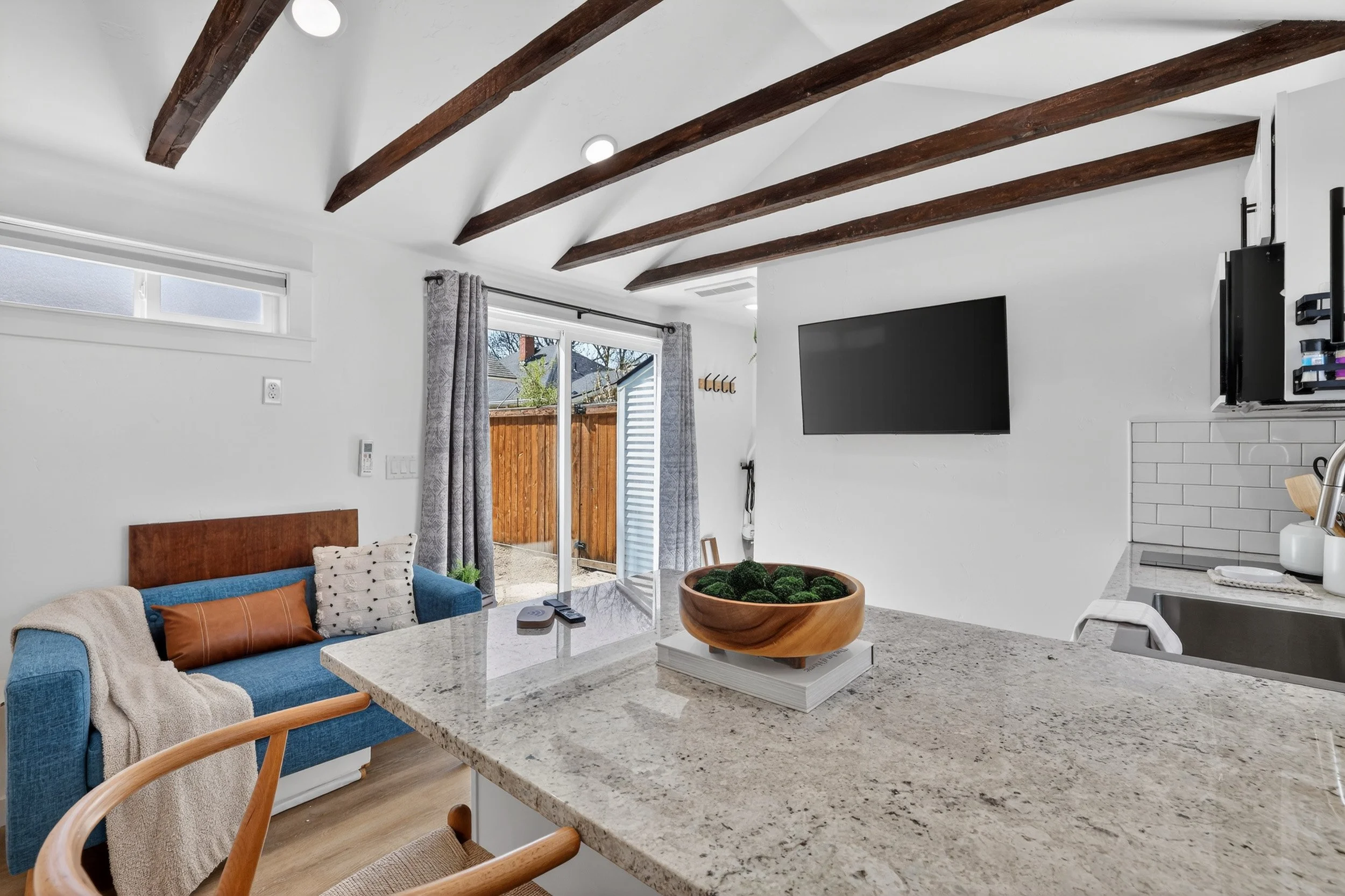 Open-concept living space with a granite kitchen island, blue sofa with pillows, sliding glass door with curtains leading outside, white walls, wooden ceiling beams, and a wall-mounted TV.