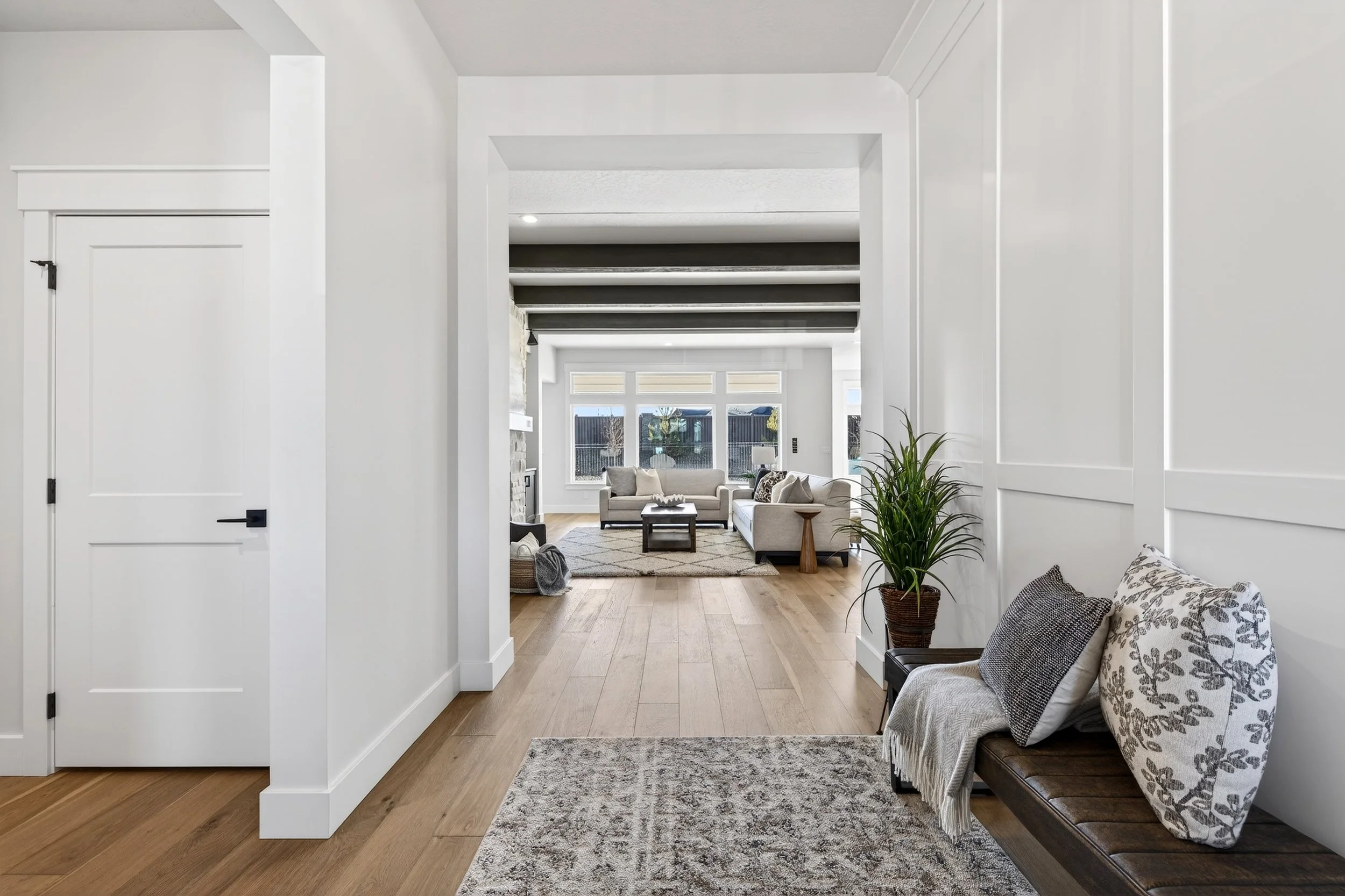View of a modern living room with beige sofas, large windows, a stone fireplace, wooden flooring, and a potted plant near a bench with pillows.