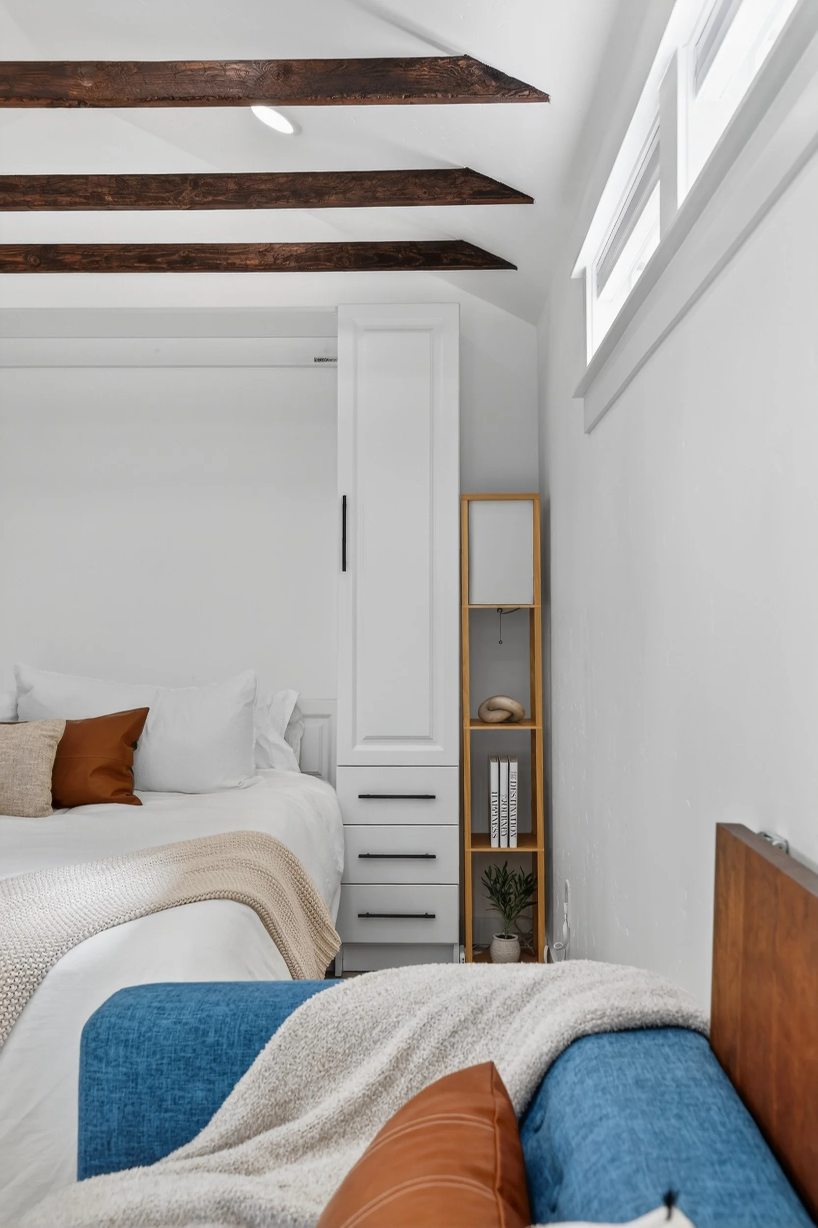 Cozy bedroom with white walls, dark wood ceiling beams, a white bed with beige and brown pillows, a blue armchair with a beige blanket, and a narrow wooden shelf with decorative items and books.