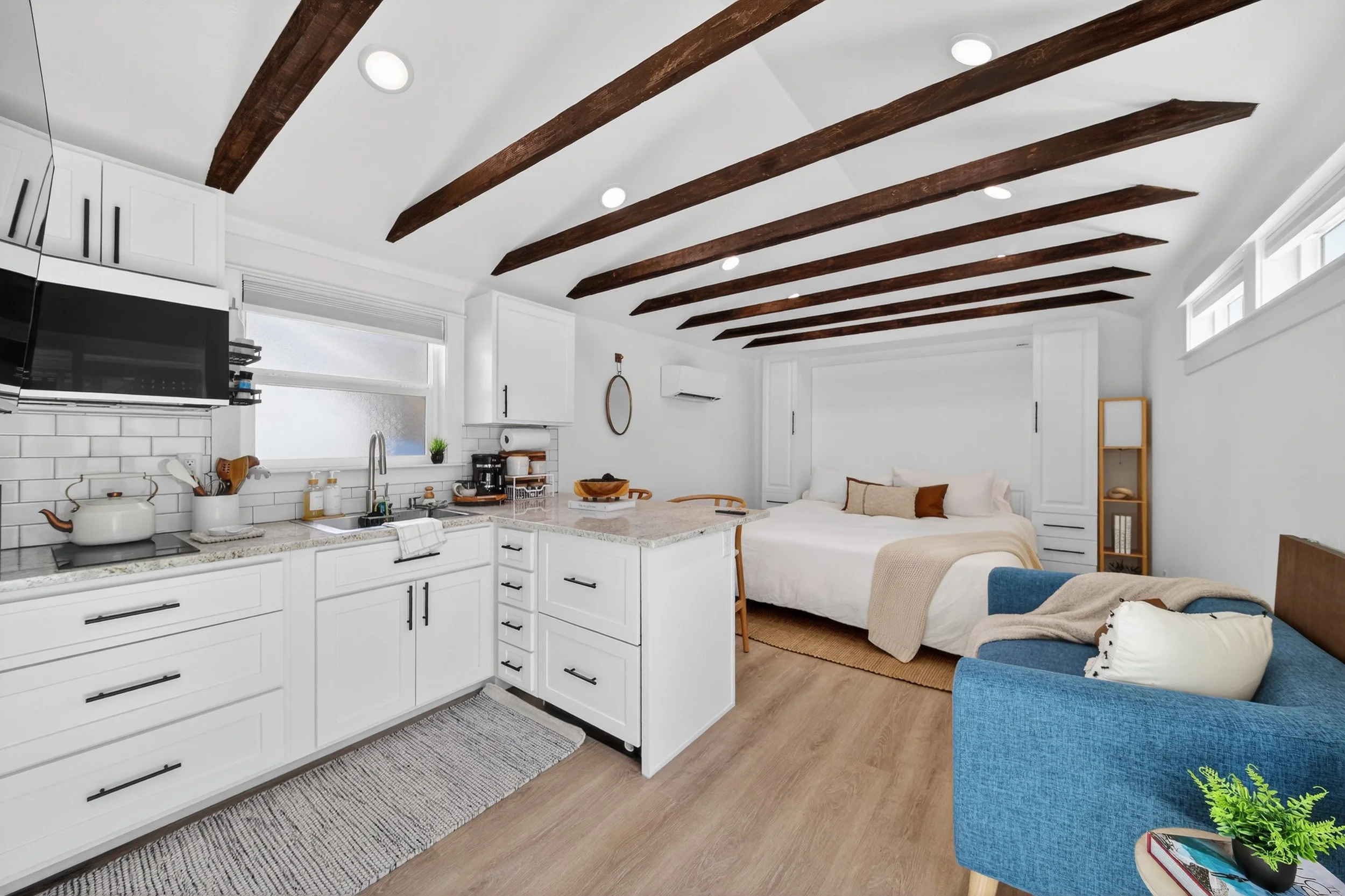 A studio apartment with a white kitchen, wooden beams on the ceiling, a bed with white linens, a blue armchair with pillows, and small windows near the ceiling.