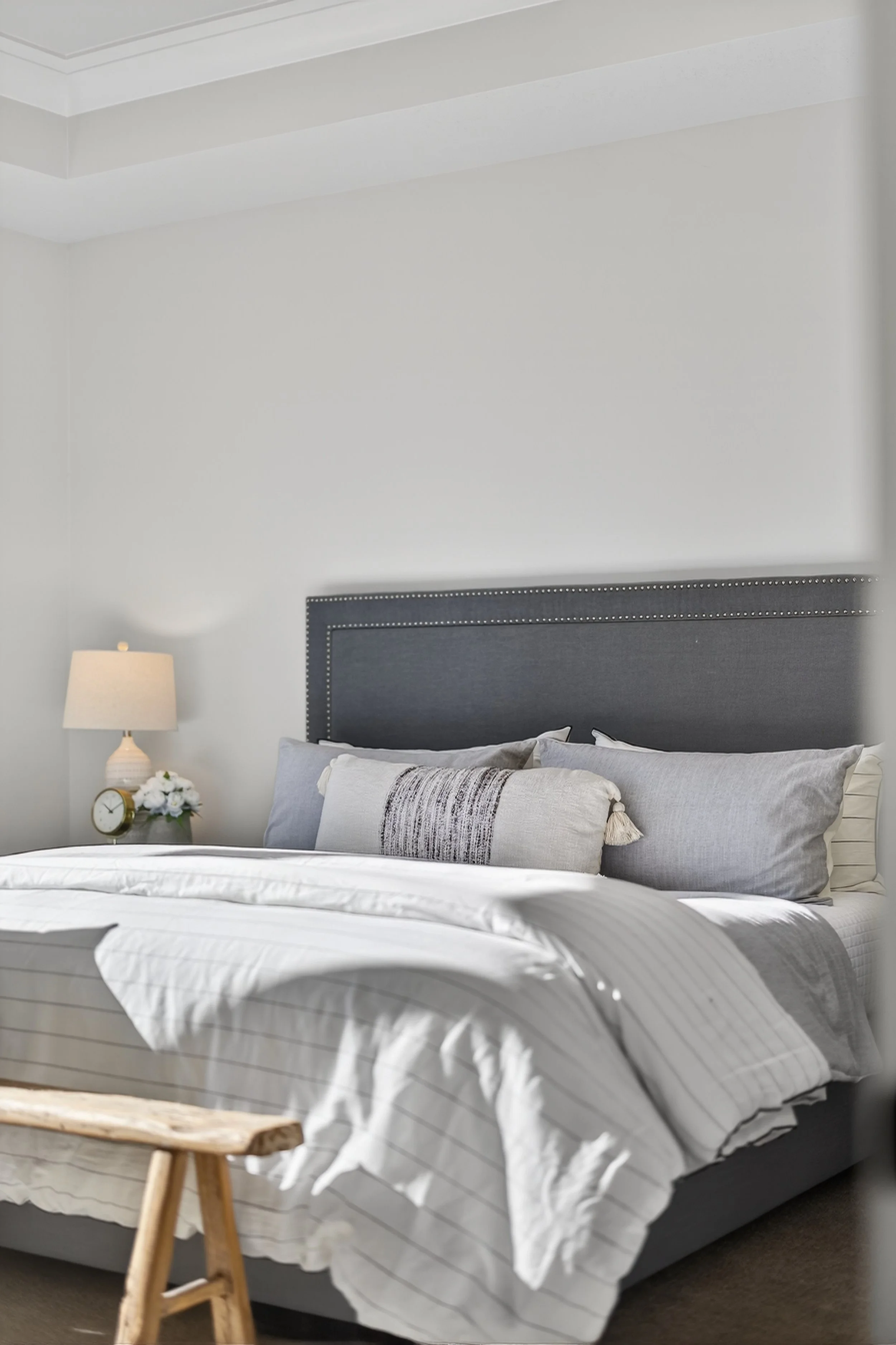 A neatly made bed with gray and white bedding, pillows, and a dark gray headboard in a minimalistic bedroom. A small bedside table with a lamp, flowers, and a clock is visible to the left.