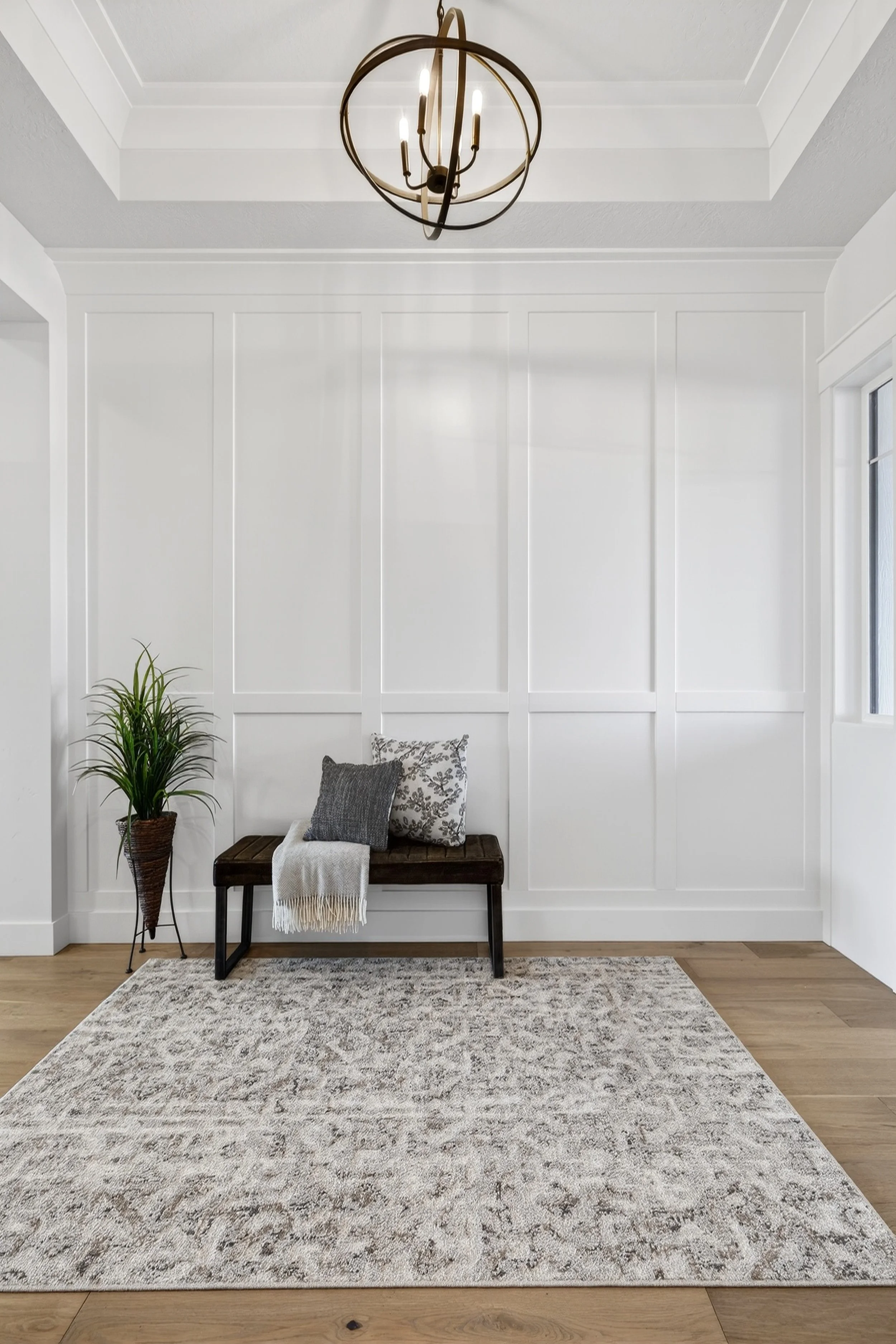 Minimalist entryway with a wooden bench, two decorative pillows, a throw blanket, a potted plant, a textured area rug, and a chandelier in a bright room with wood flooring.