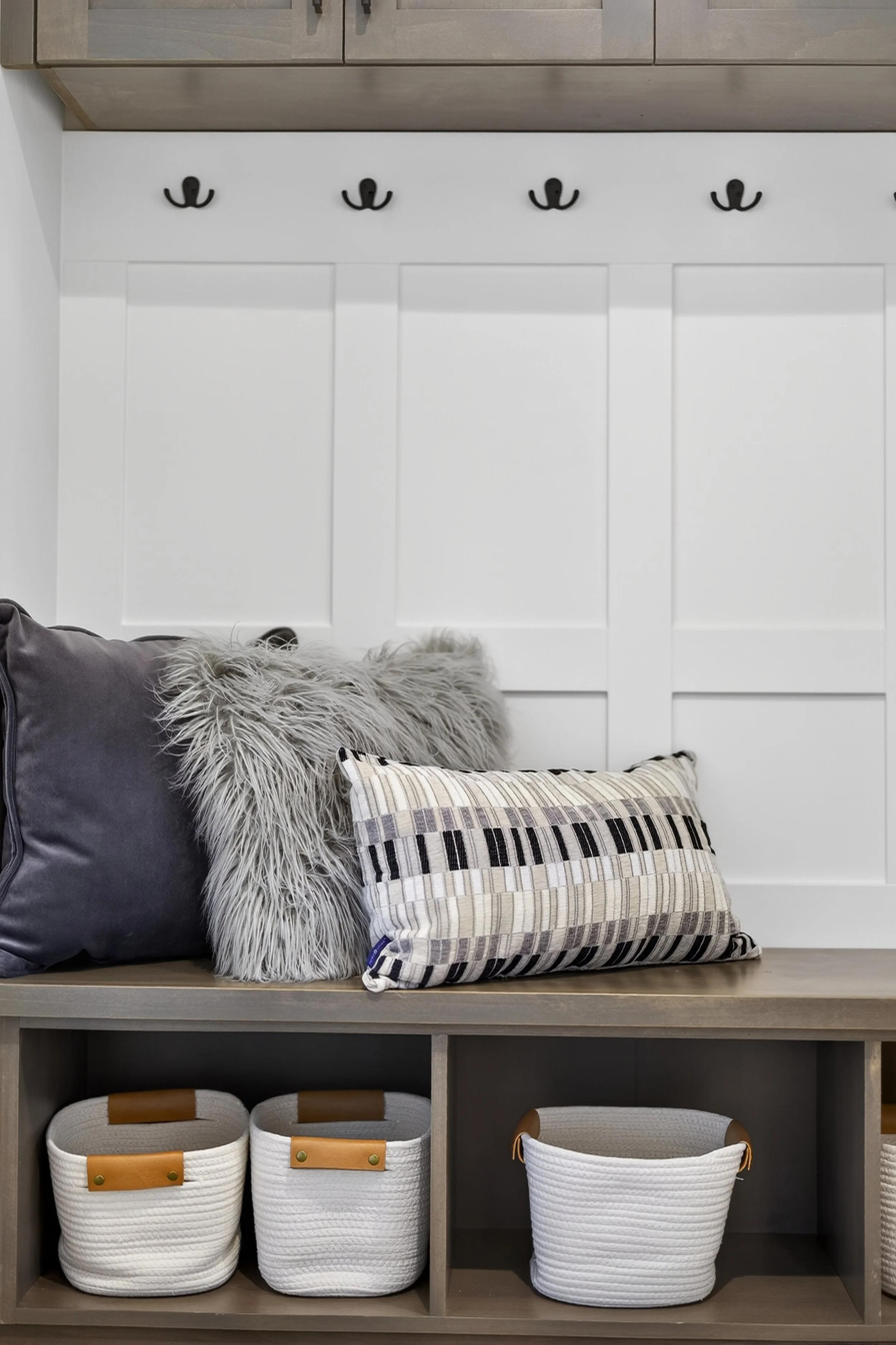 A built-in bench with decorative pillows in front of a white paneled wall with black coat hooks, and storage baskets underneath.