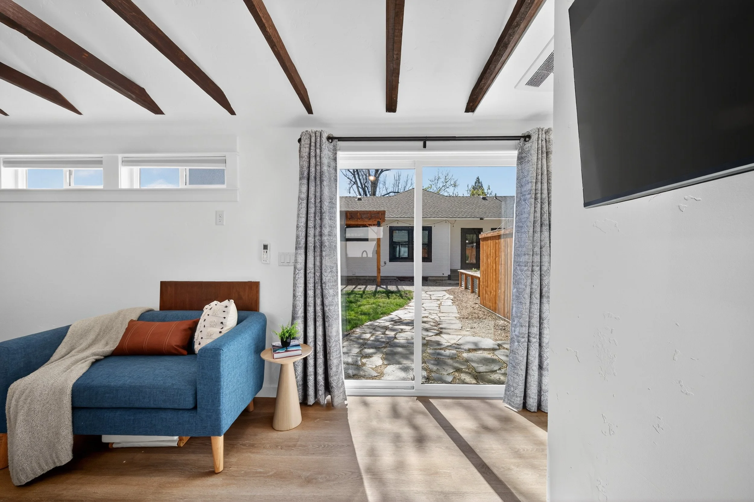Living room with sliding glass door leading to backyard, blue sofa with throw blanket and pillows, small side table with plant and books, wood-beamed ceiling, and a mounted flat-screen TV.
