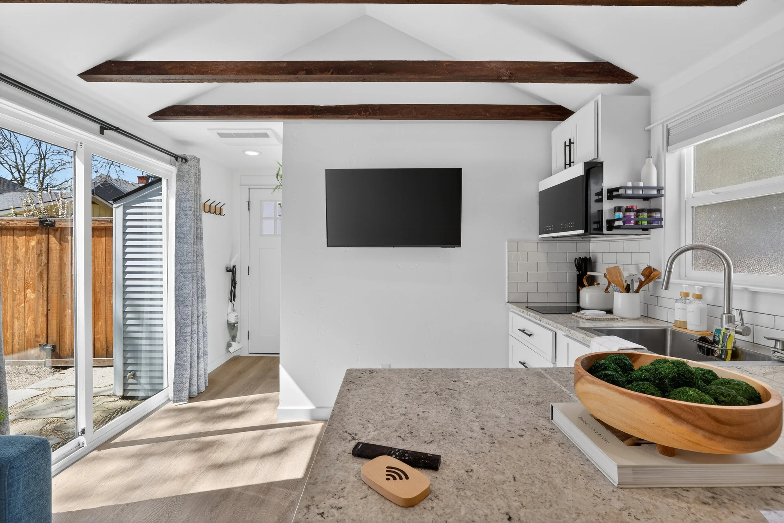 Kitchen with white cabinets, island countertop with green broccoli in wooden bowl, microwave, wall-mounted TV, sliding glass door, and wooden ceiling beams.