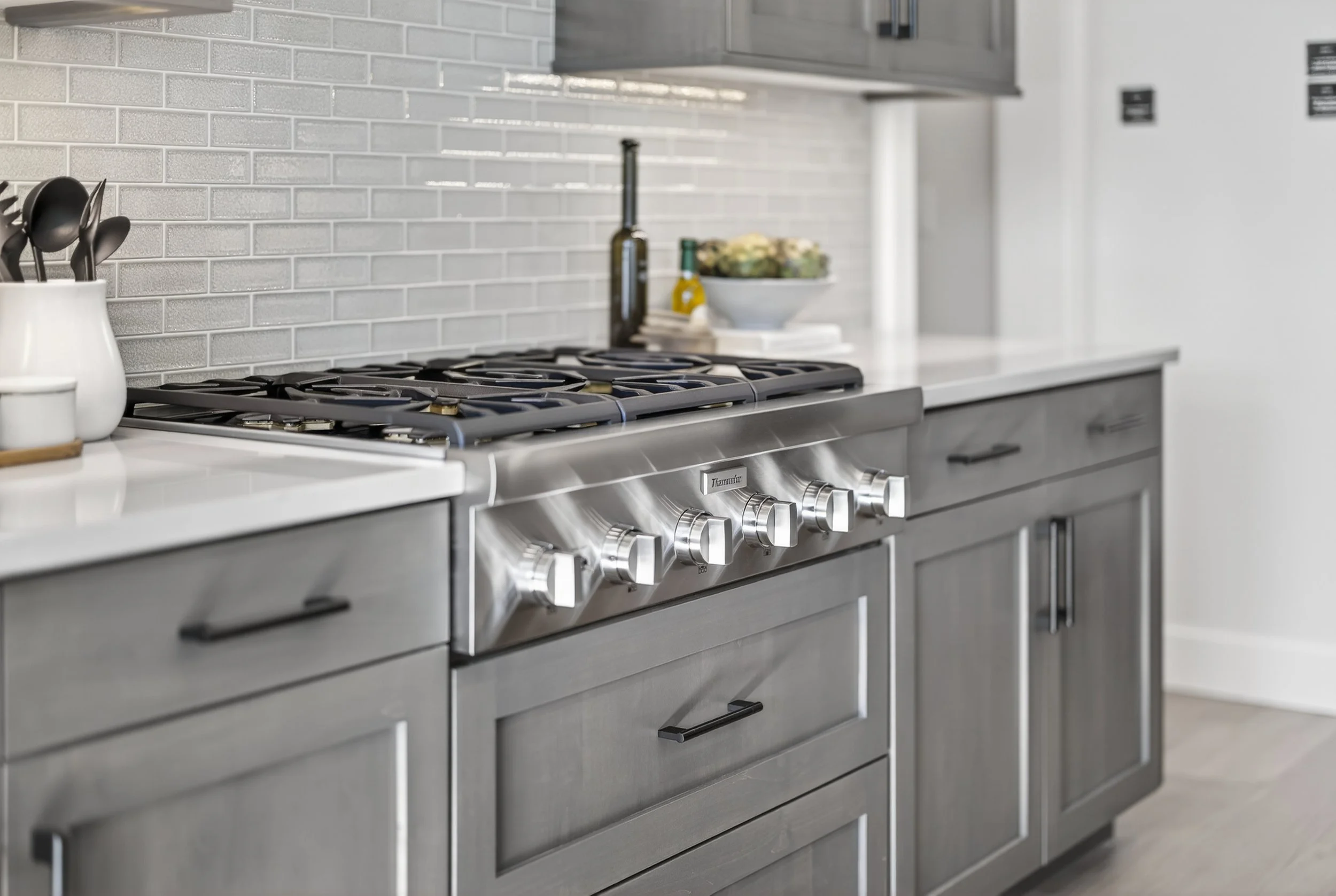 Modern kitchen with gray cabinets, a stainless steel gas stove with four burners and control knobs, white countertops, a gray brick backsplash, and various kitchen items including a dish rack with utensils, a bottle of olive oil, a bowl of vegetables