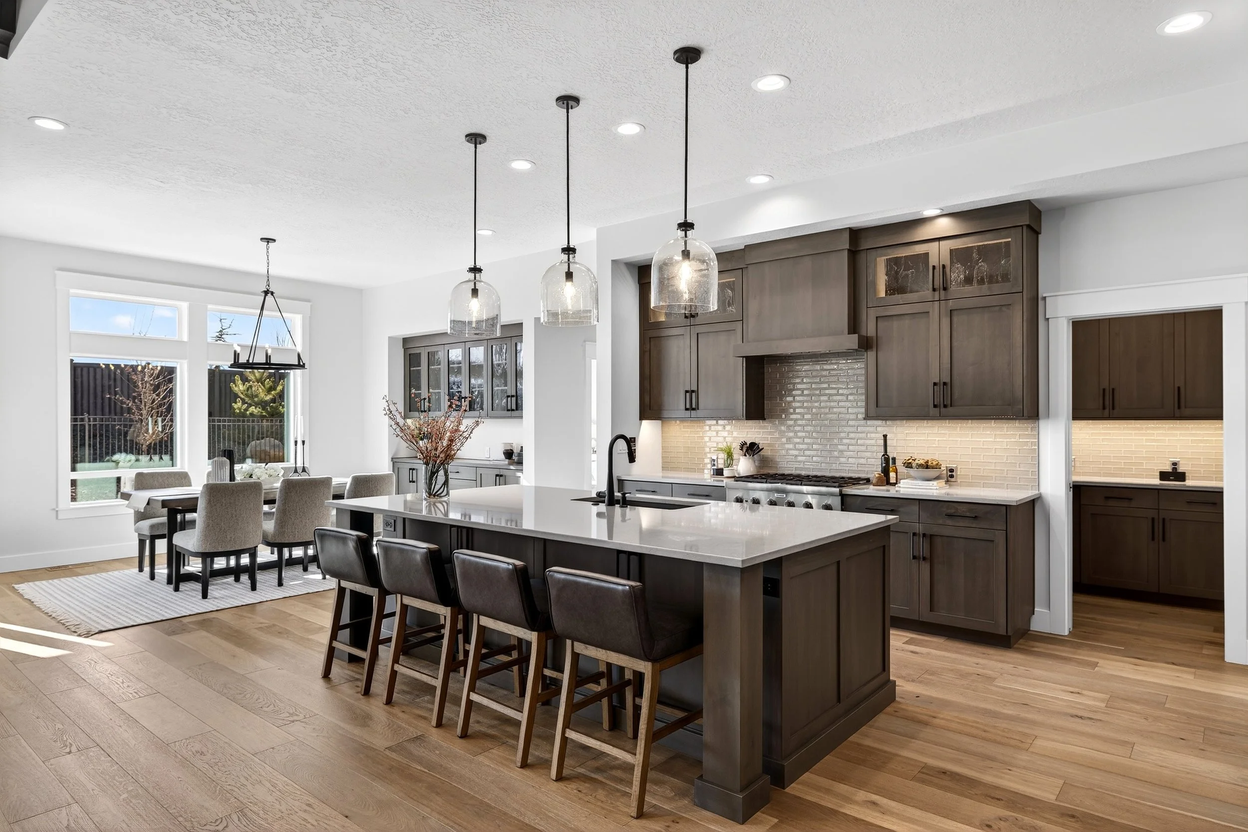 Modern kitchen with dark wood cabinets, white island with black barstools, and dining area with beige chairs and a large window.