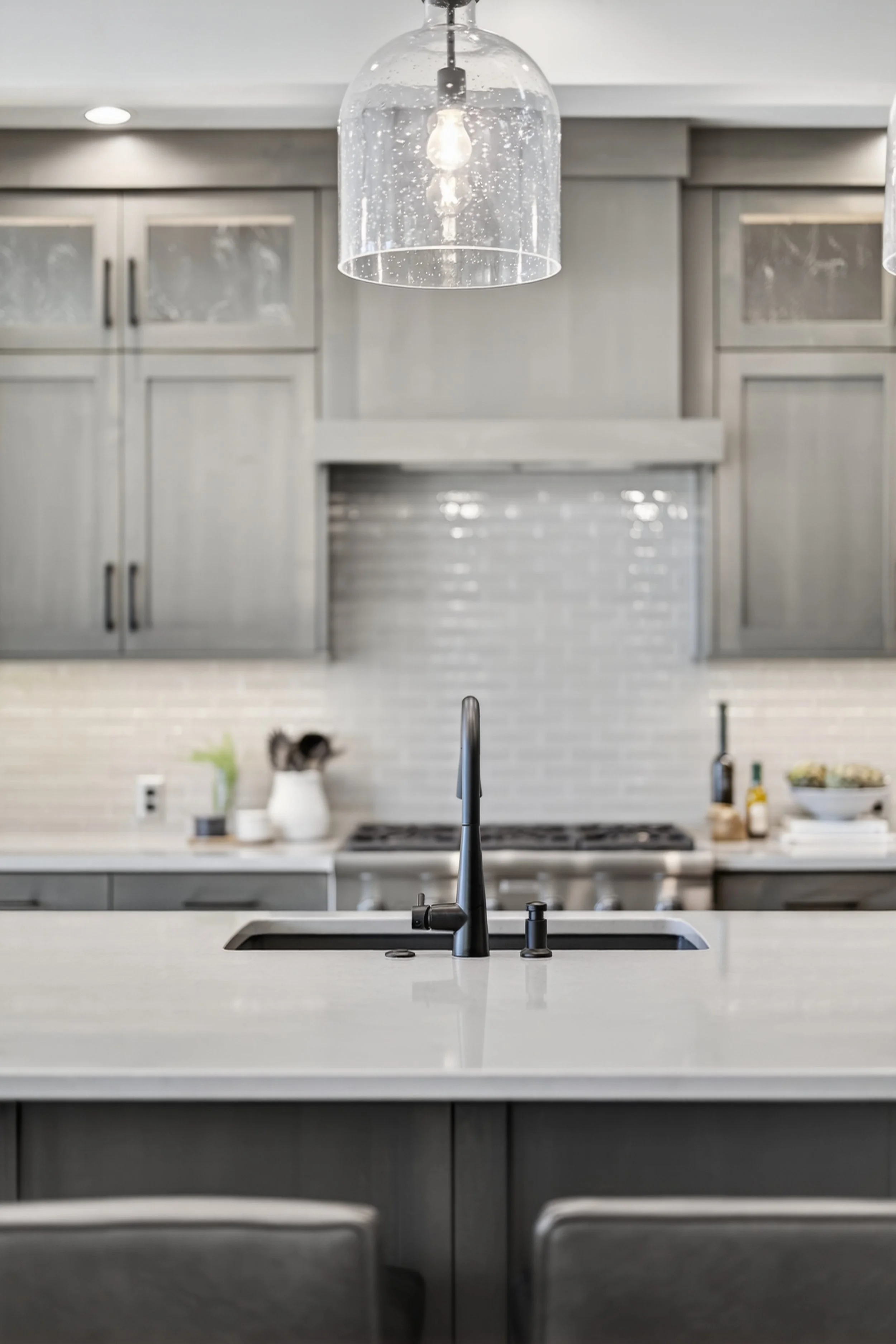 Modern kitchen with gray cabinets, a white marble island, black faucet, pendant light, and a stove in the background.