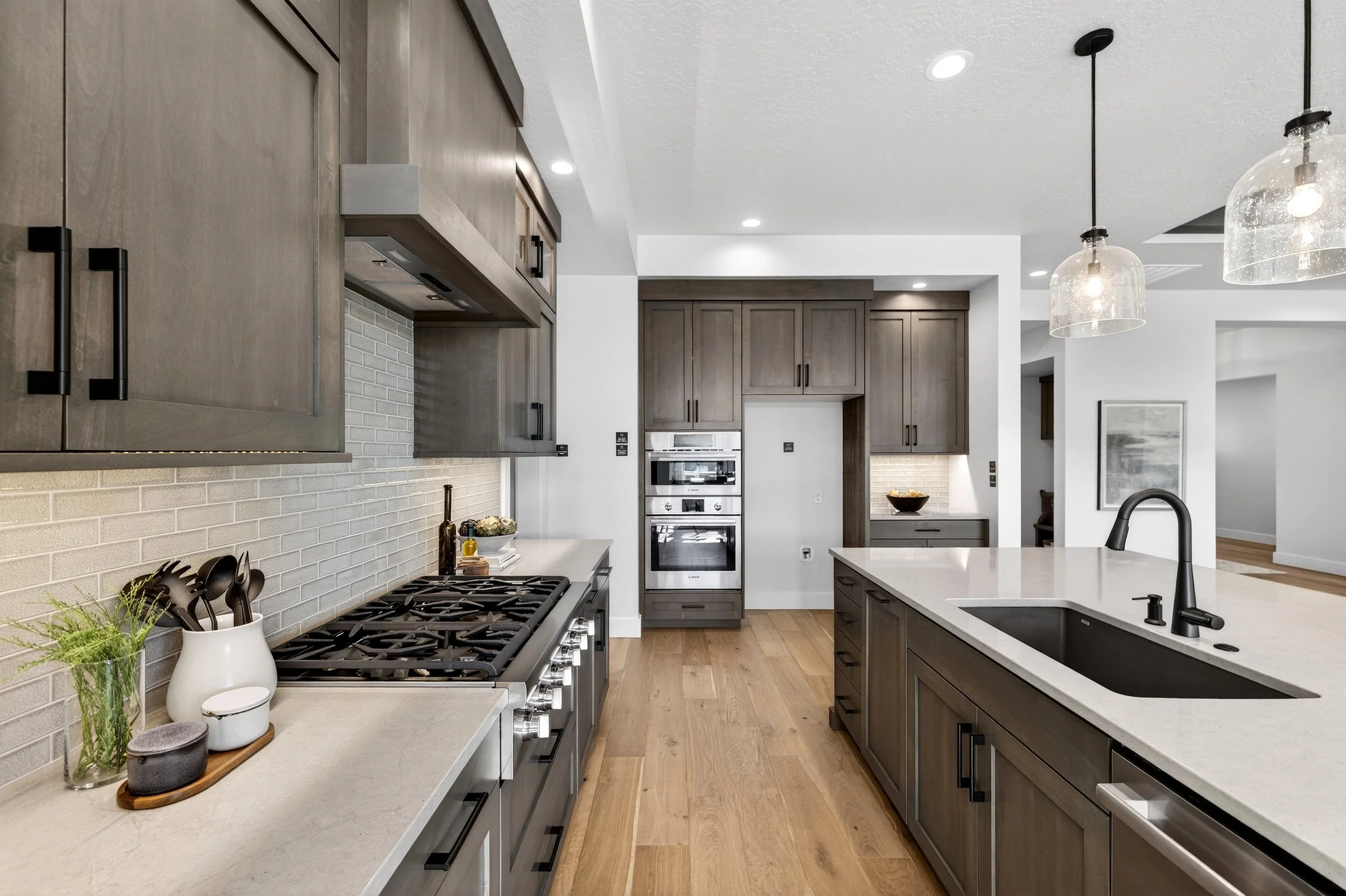 Modern kitchen with gray cabinets, white countertops, and a brick-patterned backsplash. A black stove with control knobs on the front is on the left, with kitchen utensils in a white container. In the background, stainless steel double wall ovens are