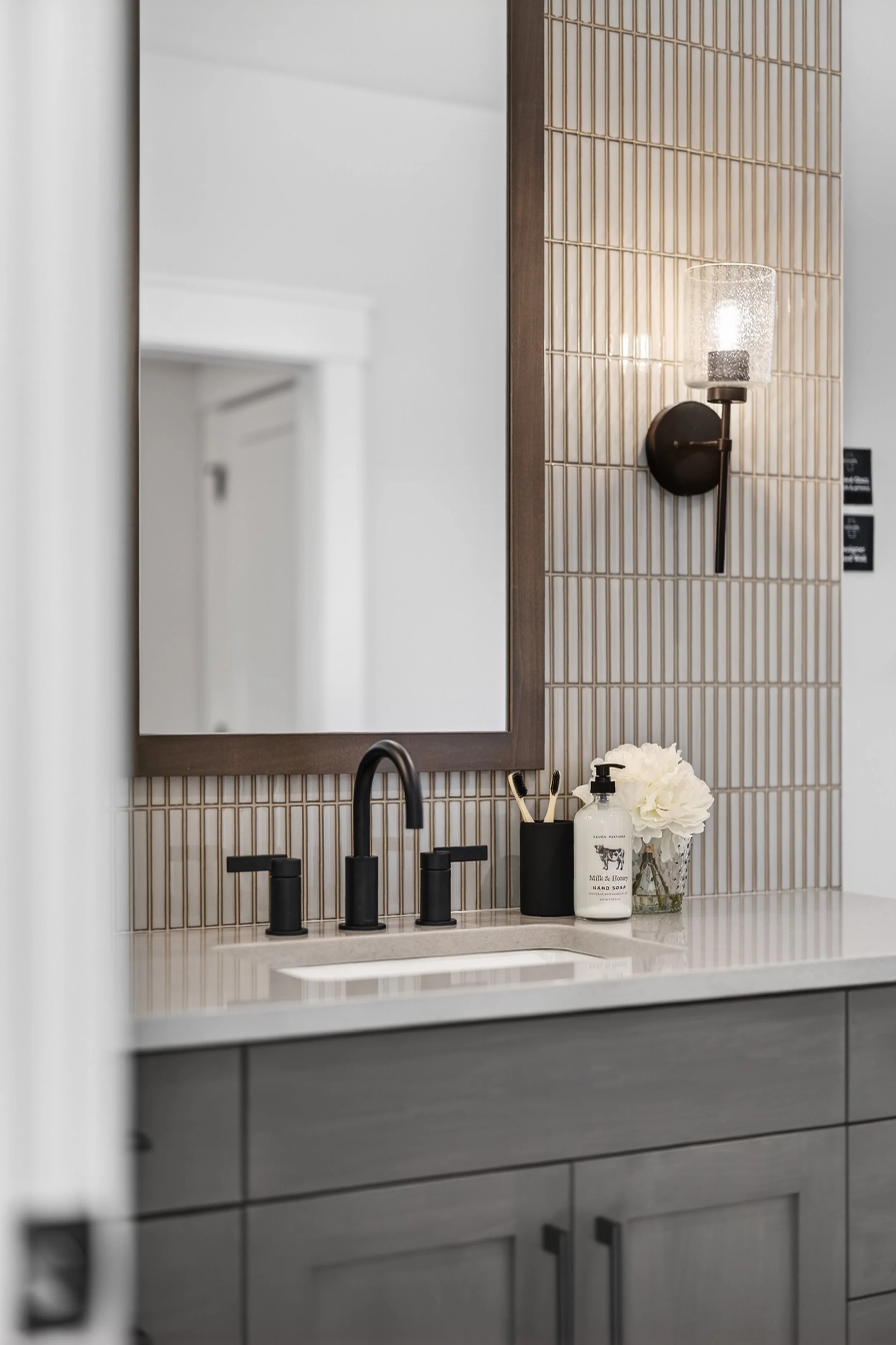 Bathroom vanity with black faucet, soap dispenser, toothbrush holder, and white flowers, against a tiled wall with a mirror and wall sconce light.