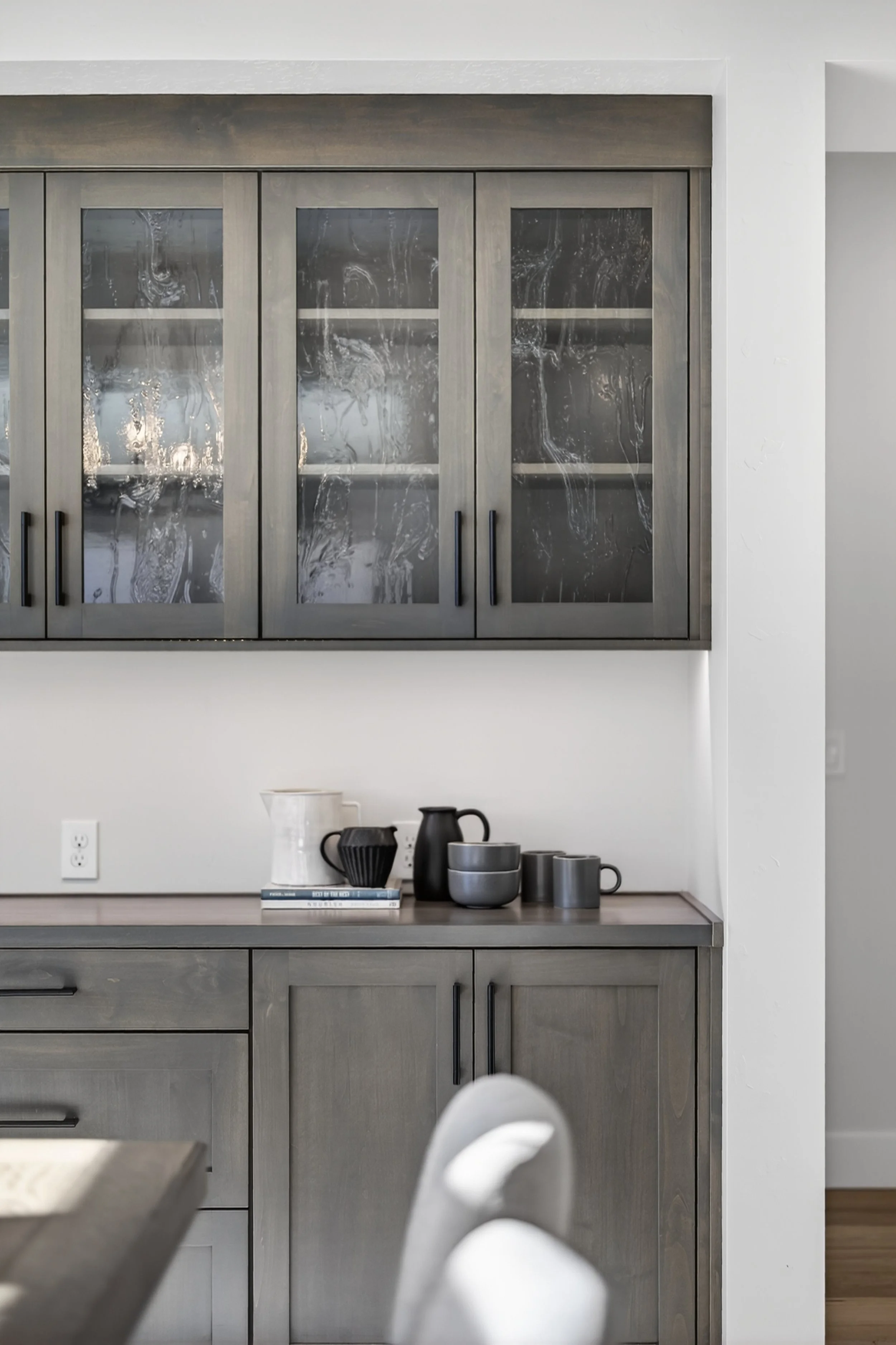 Modern kitchen with gray cabinets, glass-front upper cabinets with foggy glass, and a countertop with black and gray ceramic mugs, a bowl, and a book.