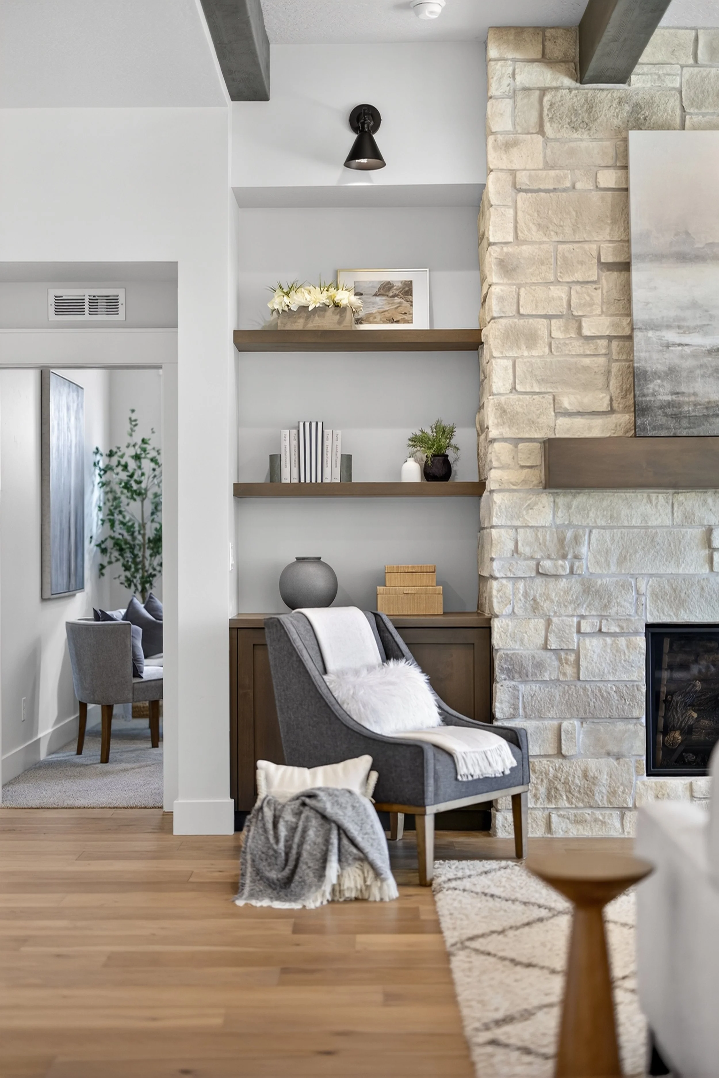 Cozy living room corner with a gray armchair, white throw blanket, furry pillow, wood side table, stone fireplace, and built-in shelves decorated with books, vases, and artwork.
