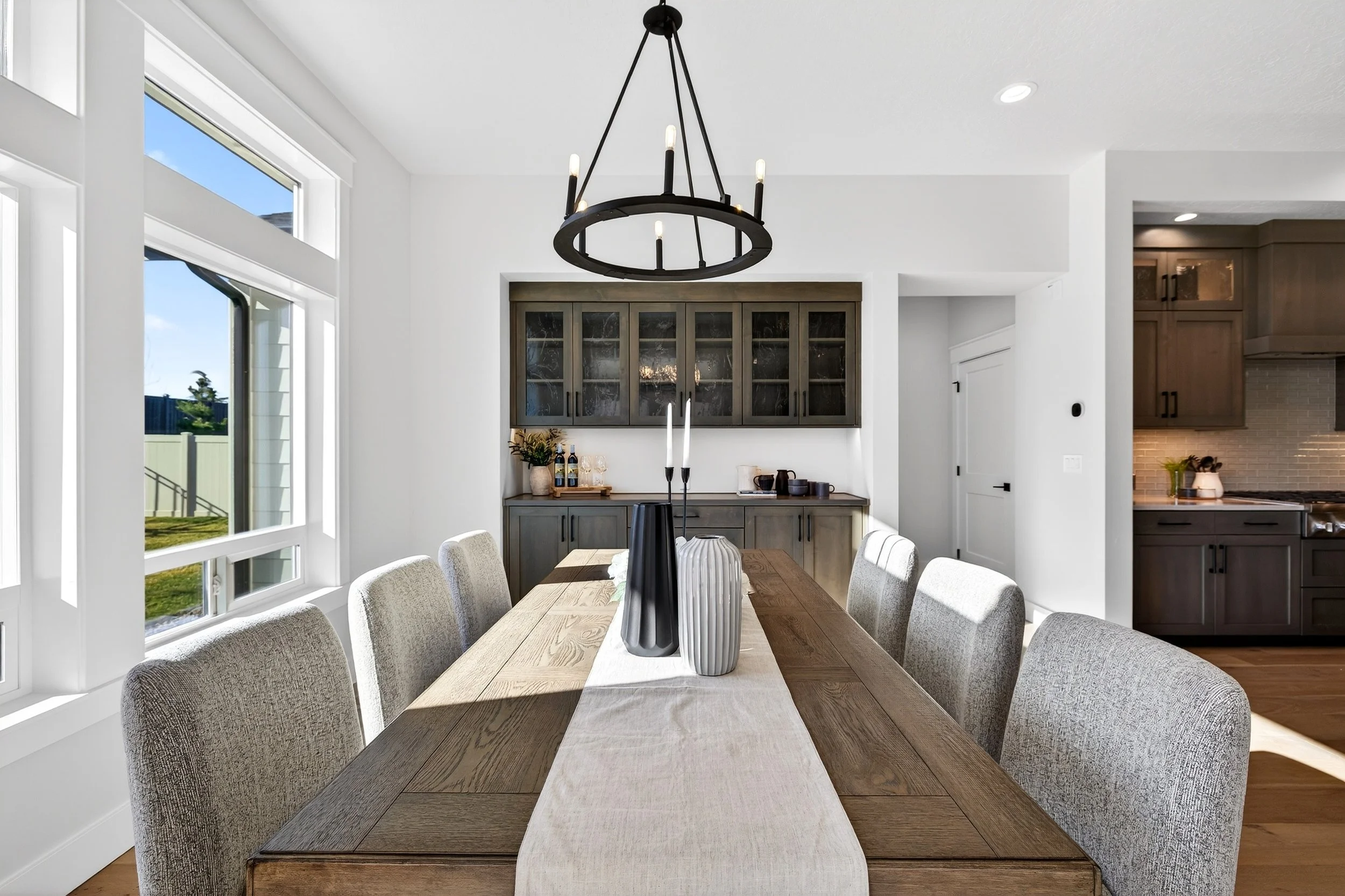 Modern dining room with wooden table, eight upholstered chairs, black chandelier, and large windows with sunlight streaming in, overlooking a backyard.