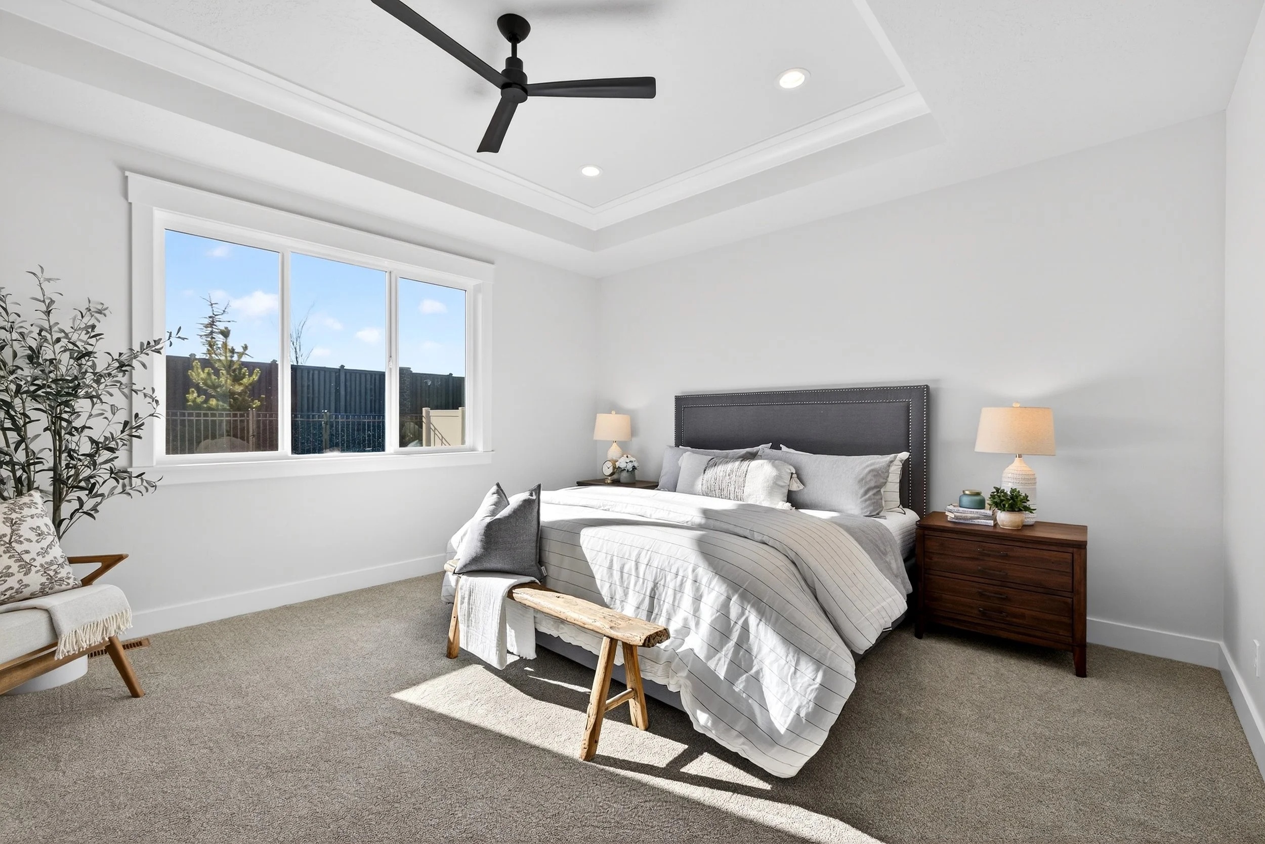 Bright bedroom with white walls, large window, gray bed with pillows, wooden nightstand with lamp, ceiling fan, and beige carpet.