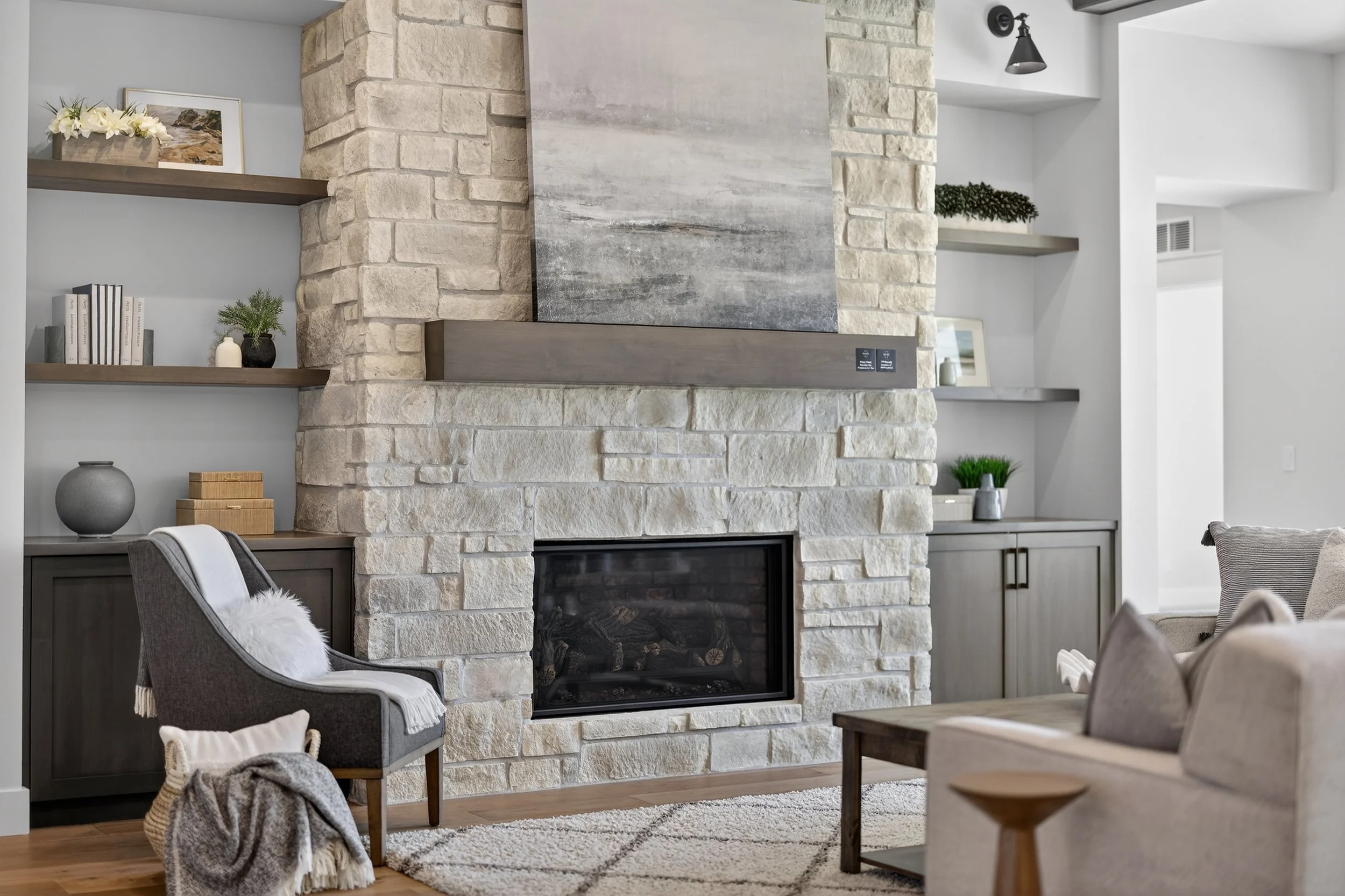Living room with a stone fireplace, built-in shelves with decor, and a gray armchair with a white throw blanket and pillow.