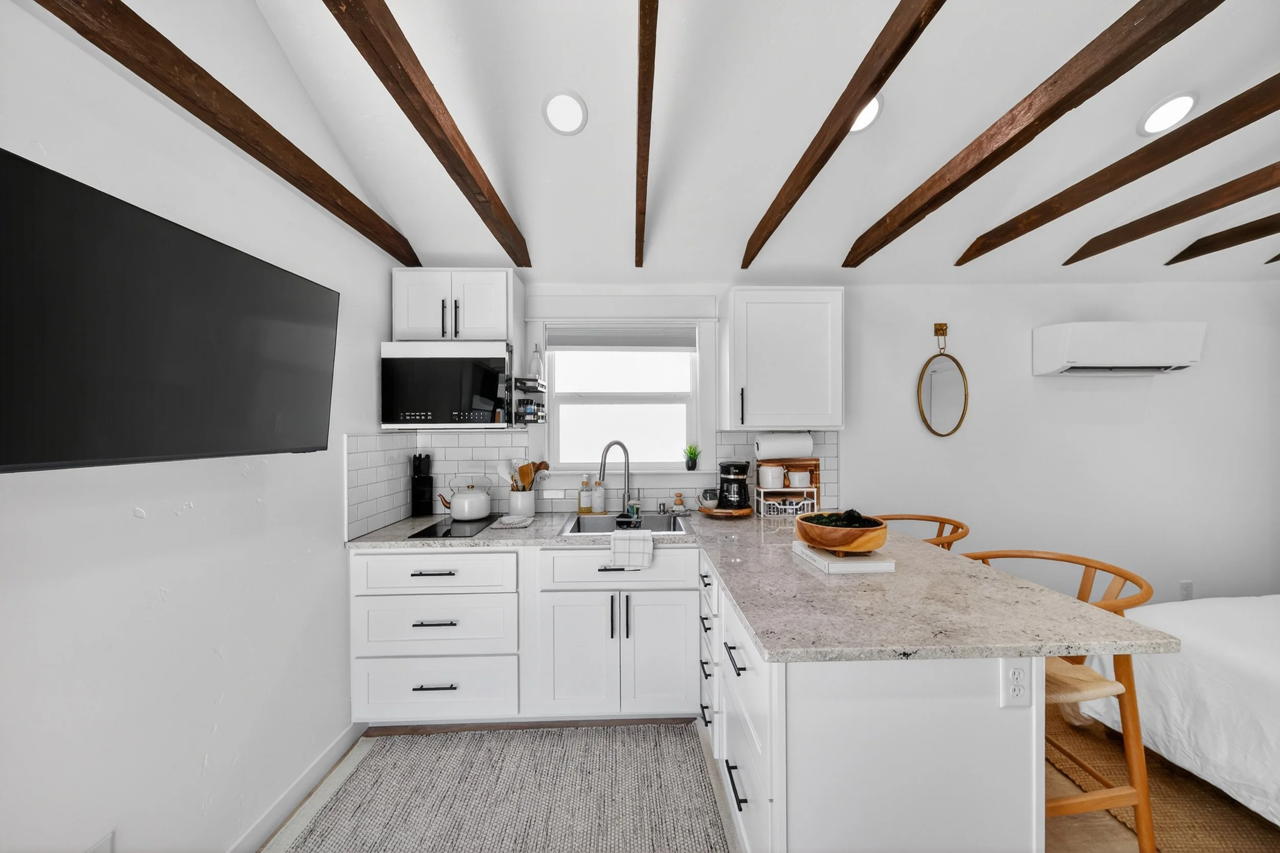 Kitchen with white cabinets, granite countertop, small window, black appliances, hanging wooden beams on white ceiling, wall-mounted TV, air conditioner, and a wooden bowl on the counter. Part of a bed and wooden chair are visible in adjacent space.