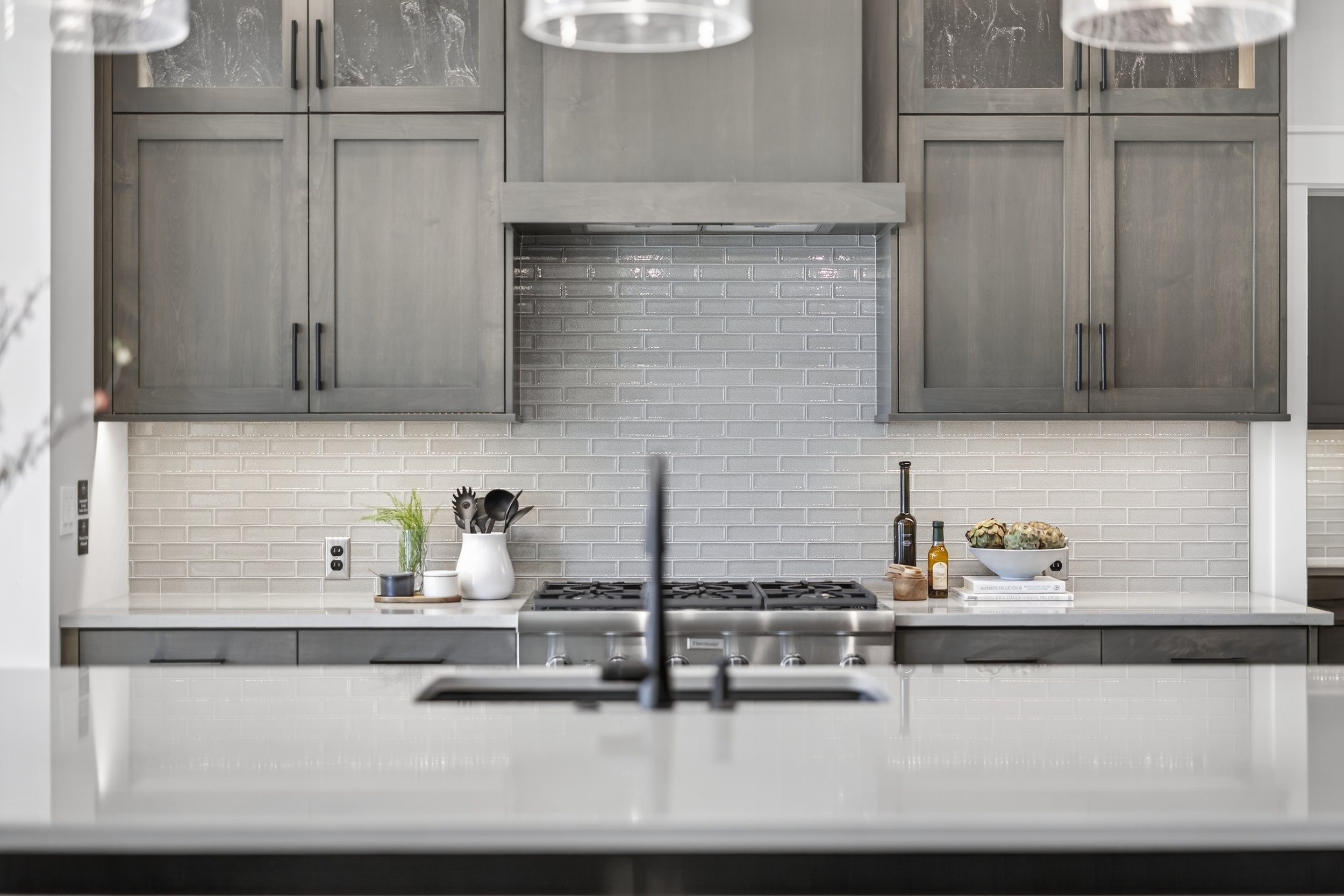Modern kitchen with gray cabinets, white backsplash tiles, and a white countertop. Visible stove, kitchen utensils, plants, and condiments on the counter.