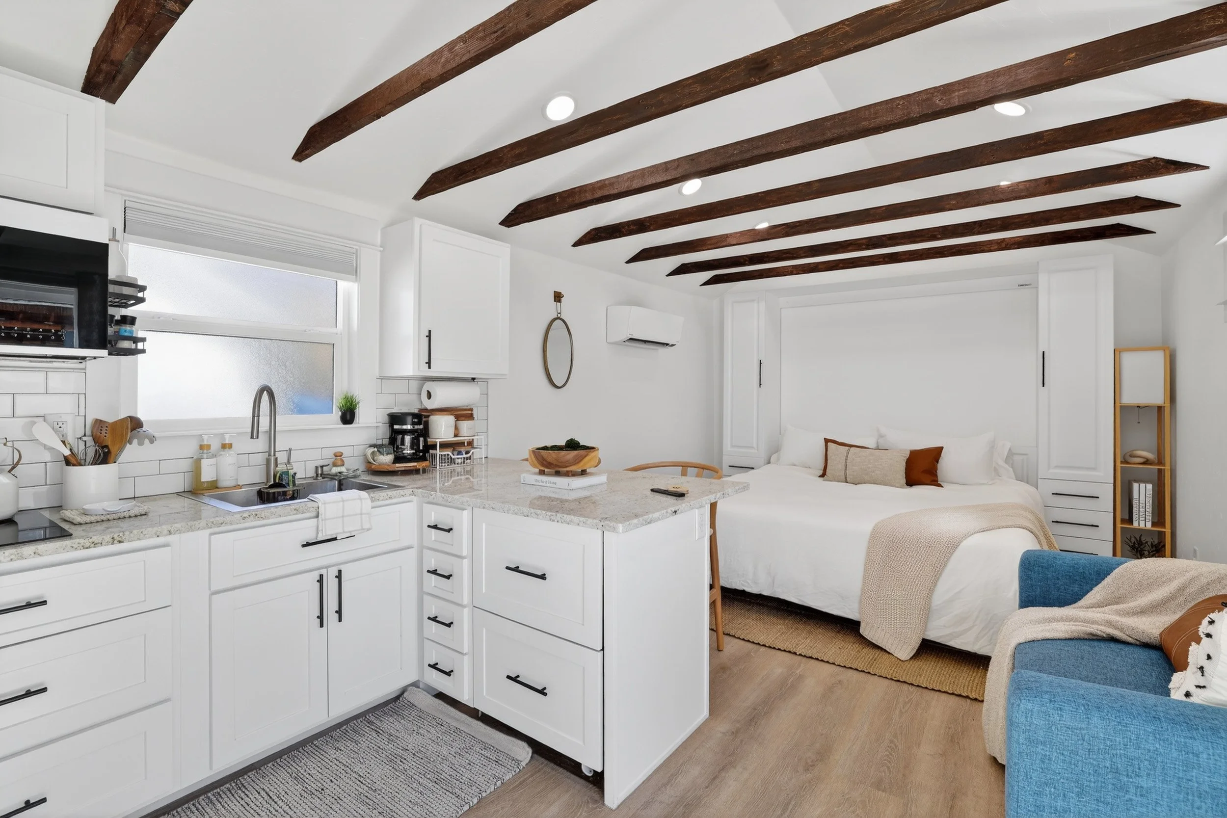 Open-concept studio apartment with kitchen and sleeping area, featuring white cabinets, granite countertops, wooden ceiling beams, a blue armchair, and a bed with neutral-colored pillows.