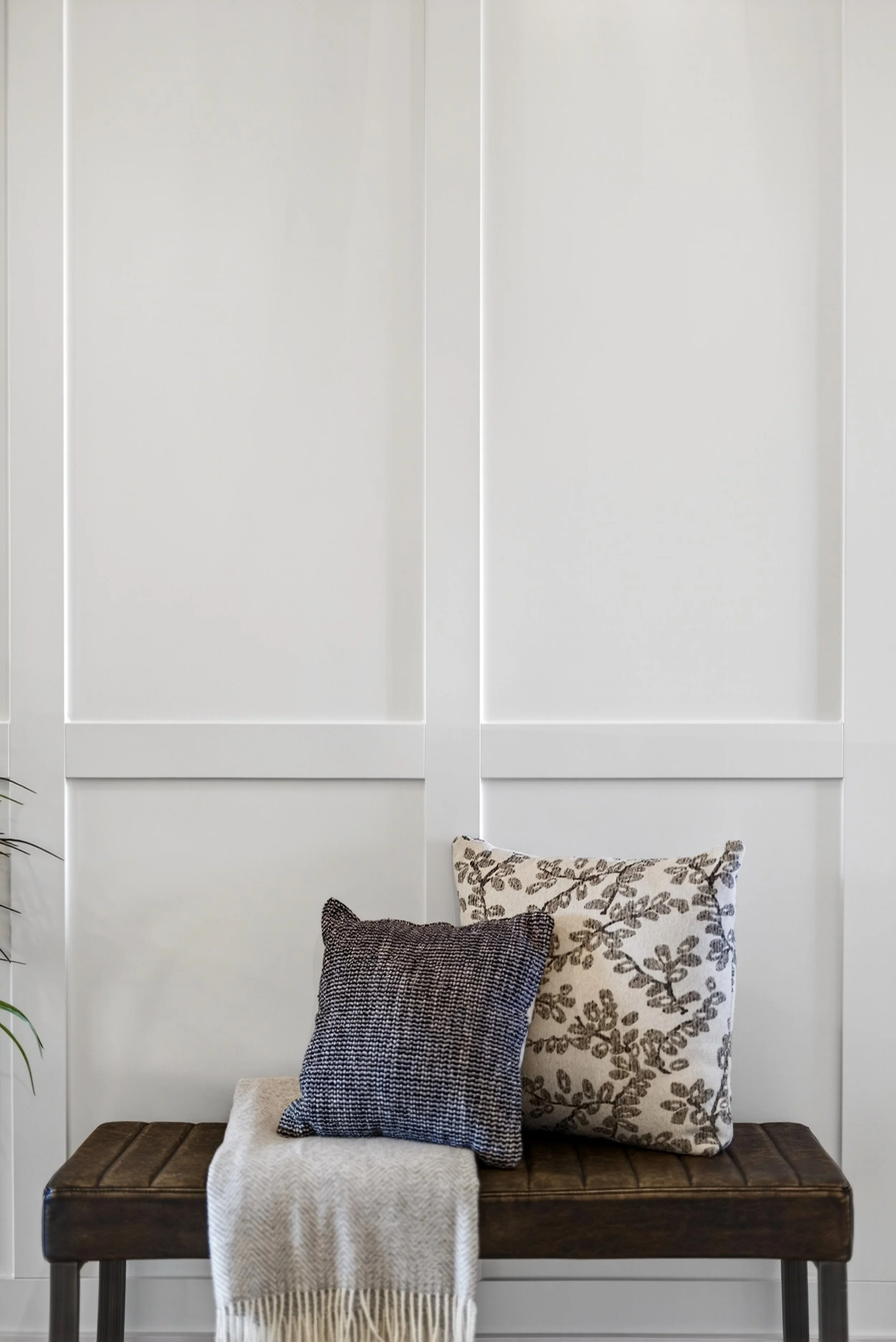 A brown leather upholstered bench with two patterned pillows and a beige throw blanket, placed against a white paneled wall.