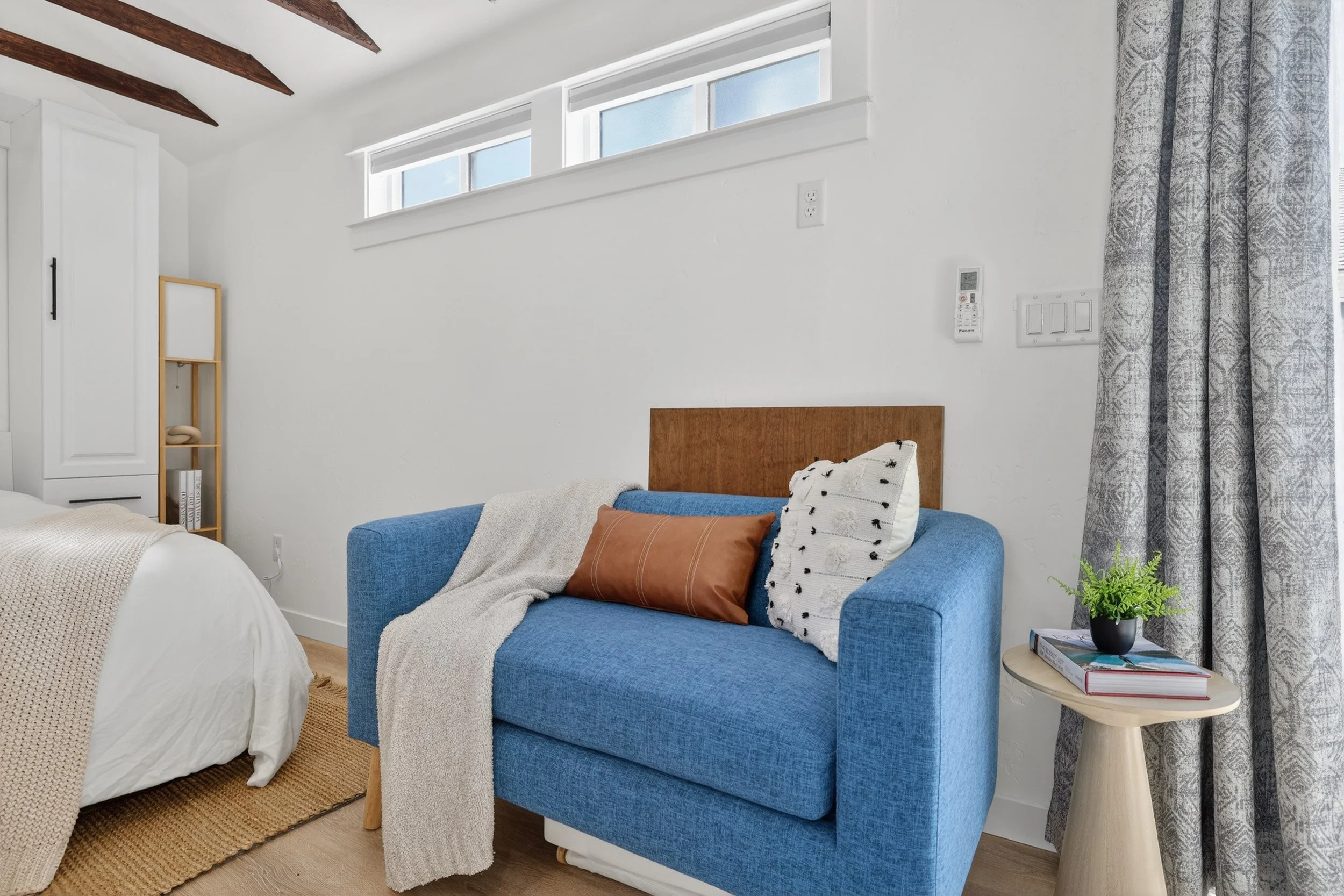 Bright bedroom corner with a blue sofa, a bed partially visible on the left, a small wooden side table with books and a plant, white walls, and a window with patterned curtains.
