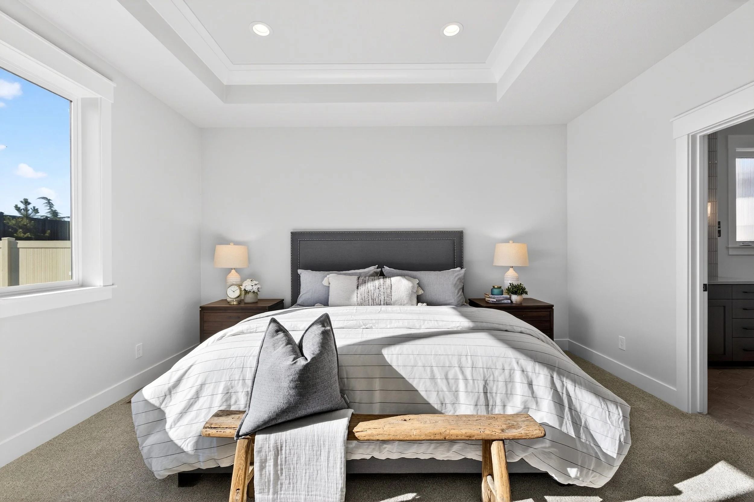 Bright bedroom with white walls and ceiling, dark gray headboard, bedside tables with lamps, and a bed with white and gray bedding and pillows. A wooden stool at the foot of the bed and a window showing a sunny outdoor scene.