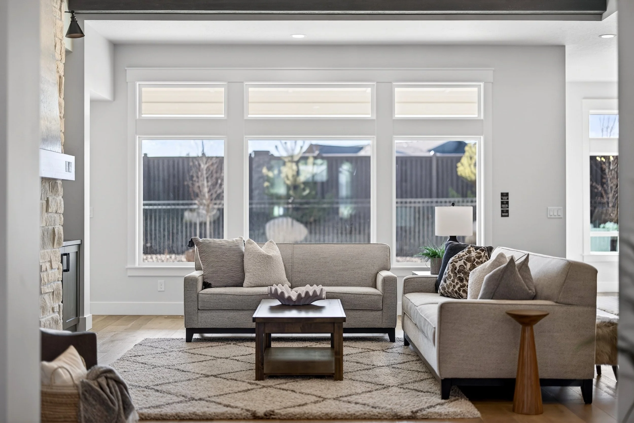 Living room with large windows, beige sofas with pillows, a wooden coffee table with a decorative bowl, a side table with a lamp and plant, and a patterned rug.