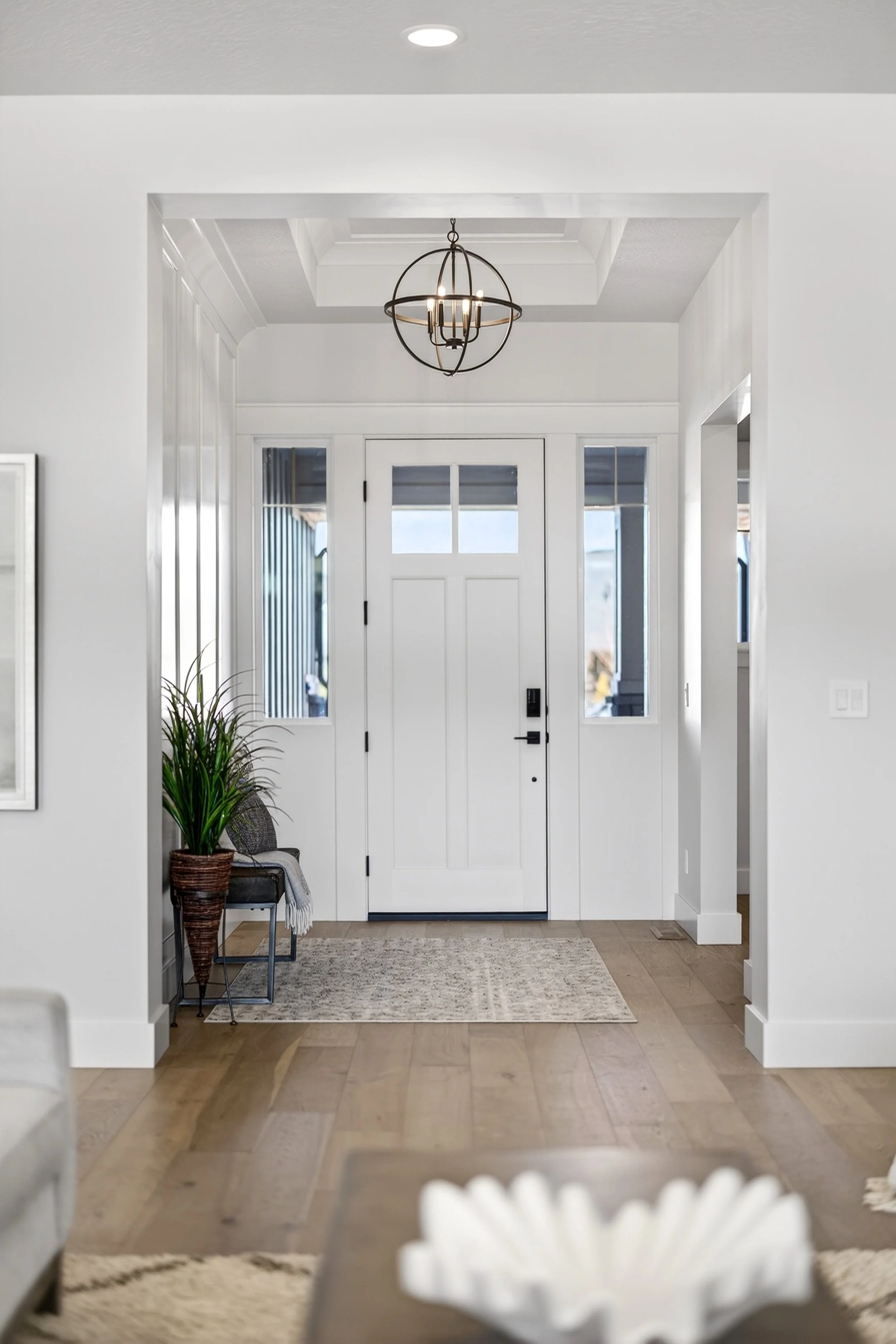 Modern white front door with glass panes, flanked by windows, in a bright entryway with wooden flooring, a rug, a potted plant, and a chandelier overhead.