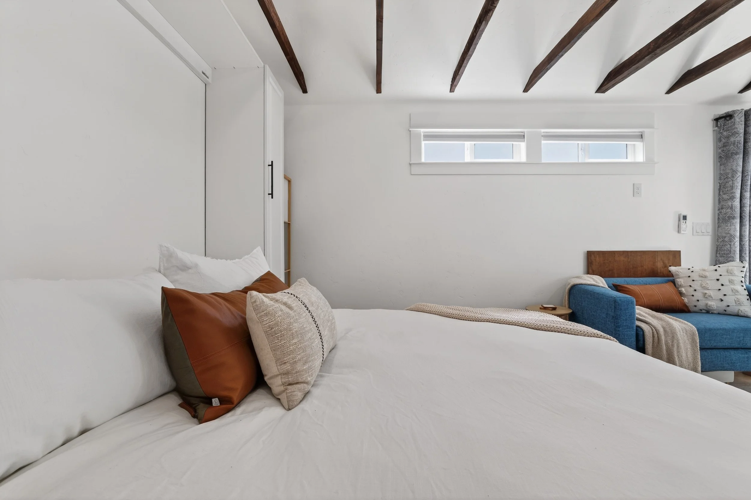 A bedroom with white walls, wooden ceiling beams, a white bed with pillows, and a blue sofa with cushions.