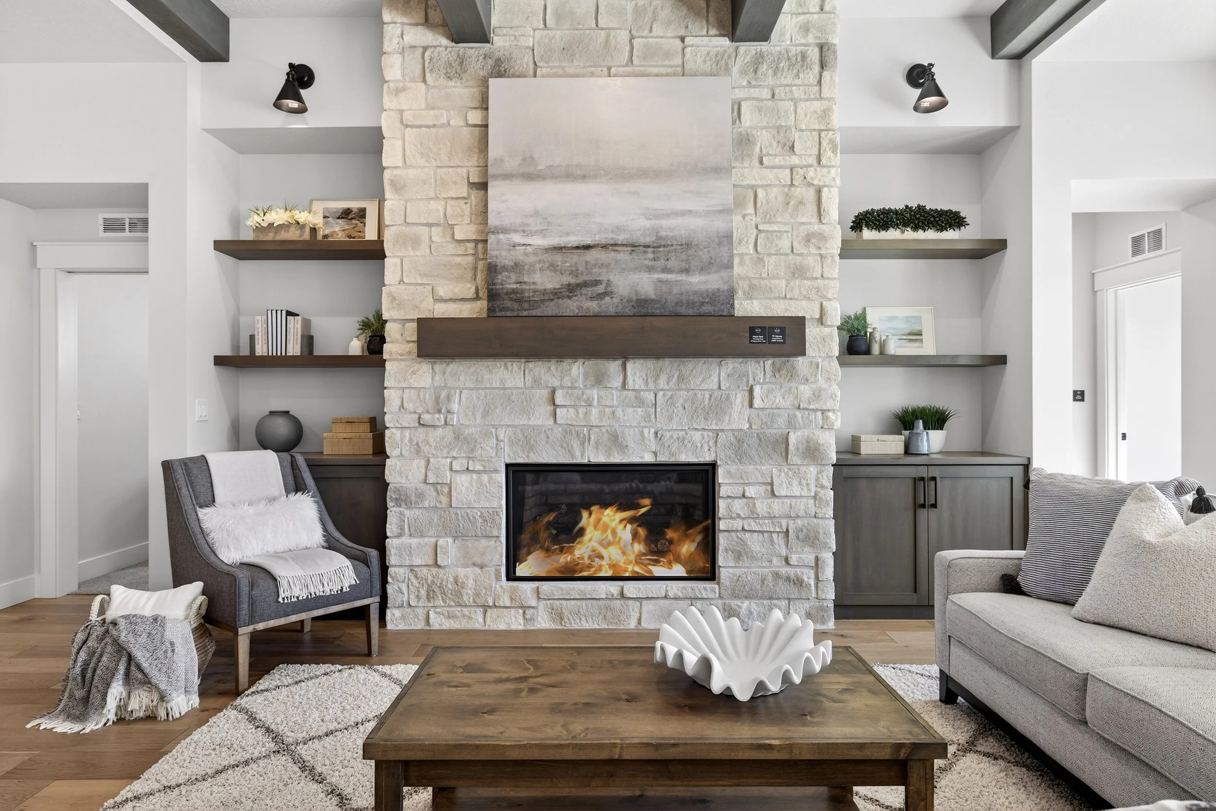 Living room with a stone fireplace, a gray armchair with a white throw blanket, a gray sofa with pillows, a wooden coffee table with a white decorative bowl, built-in shelves with decorations, and hardwood floors.