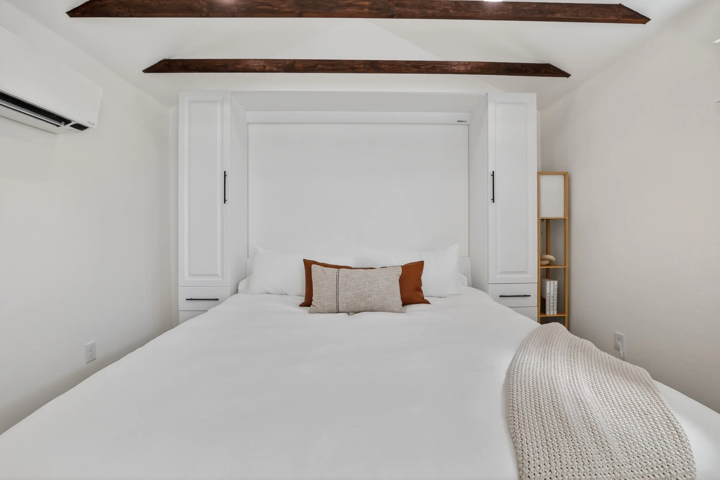 A neatly made white bed with three decorative pillows, on a white bedspread. Behind the bed is a white headboard with built-in cabinetry. To the right is a small wooden bookshelf with a phone and books. The room has white walls, dark wooden ceiling b
