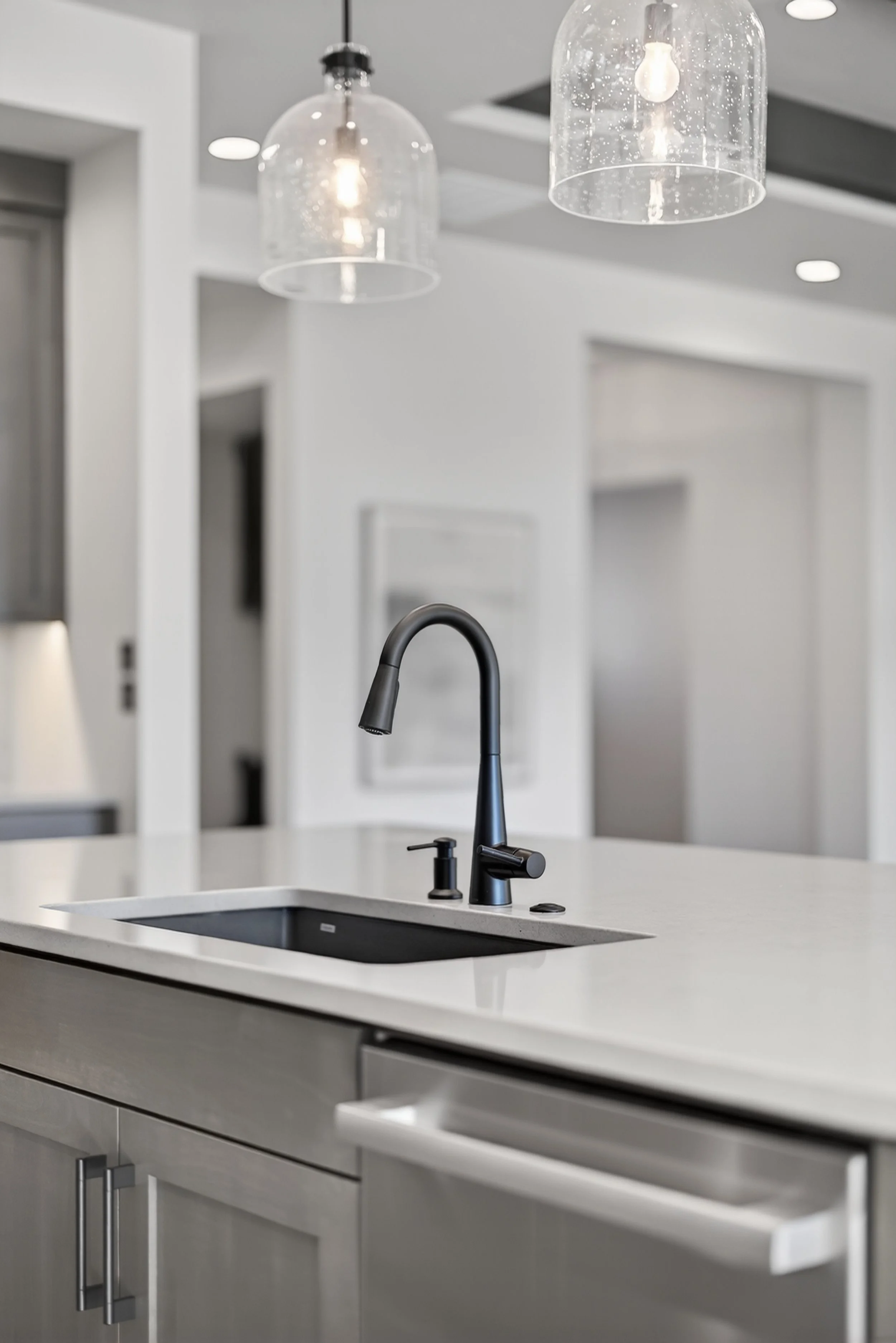 Modern kitchen with black faucet and white countertop, pendant lights overhead, and minimalist decor.