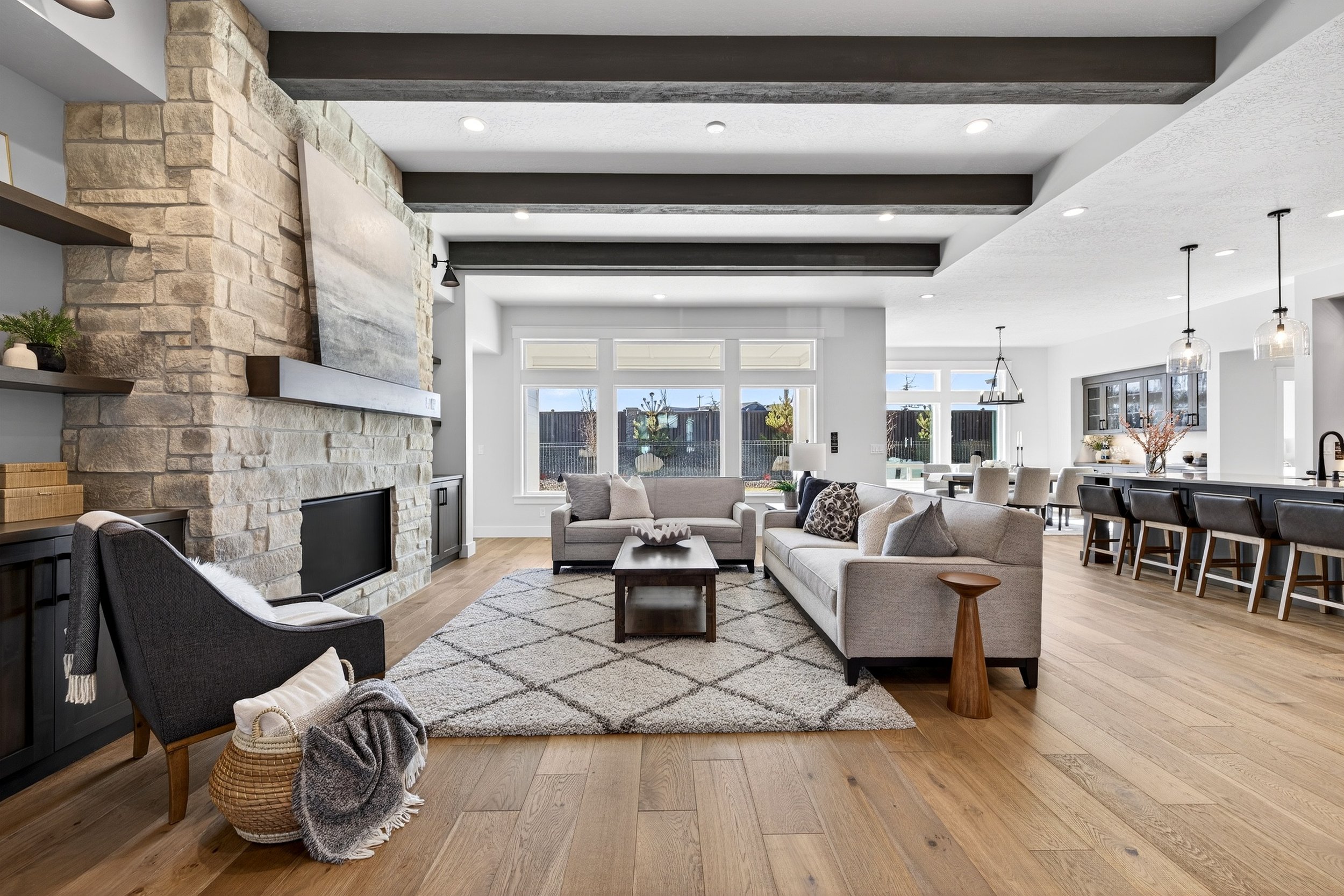 Open-concept living room with light gray sofas, wooden coffee table, stone fireplace, large windows, and hardwood flooring, with views of the outdoor backyard.