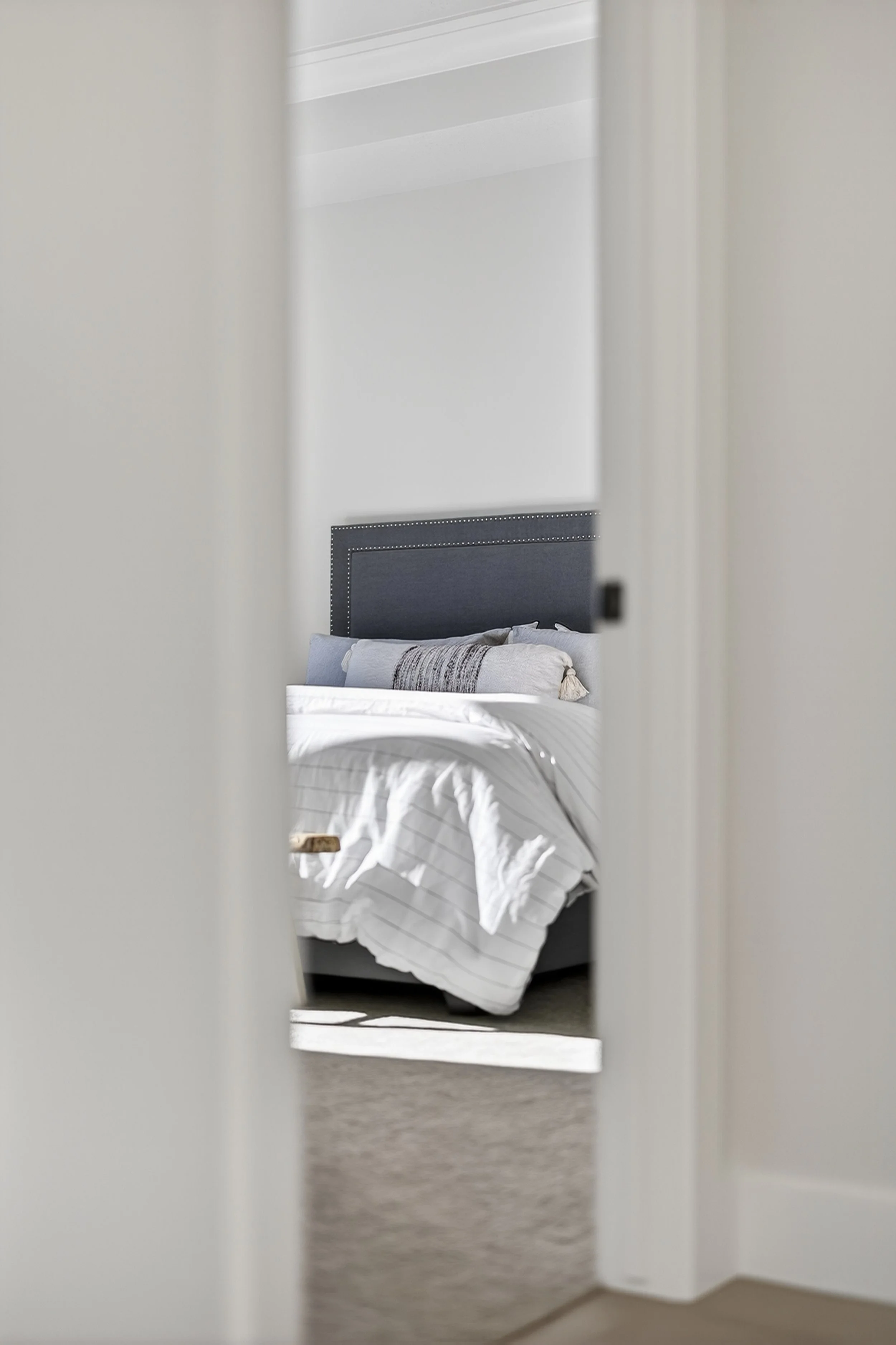 A neatly made bed with white bedding and gray pillows, seen through a doorway.