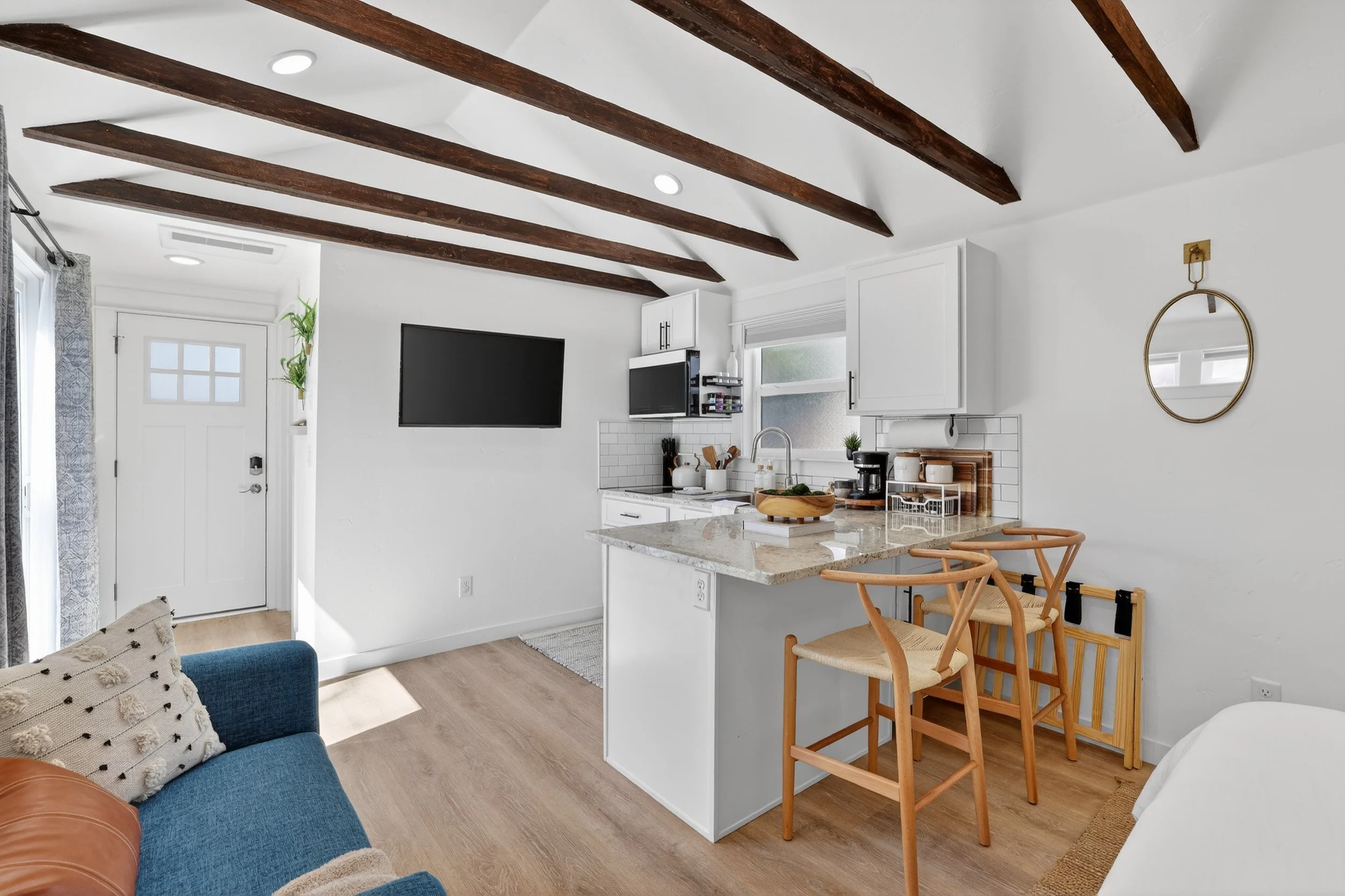 Modern kitchen and living space with white walls, wooden beams on the ceiling, a small countertop with chairs, a TV on the wall, and a door near the window.