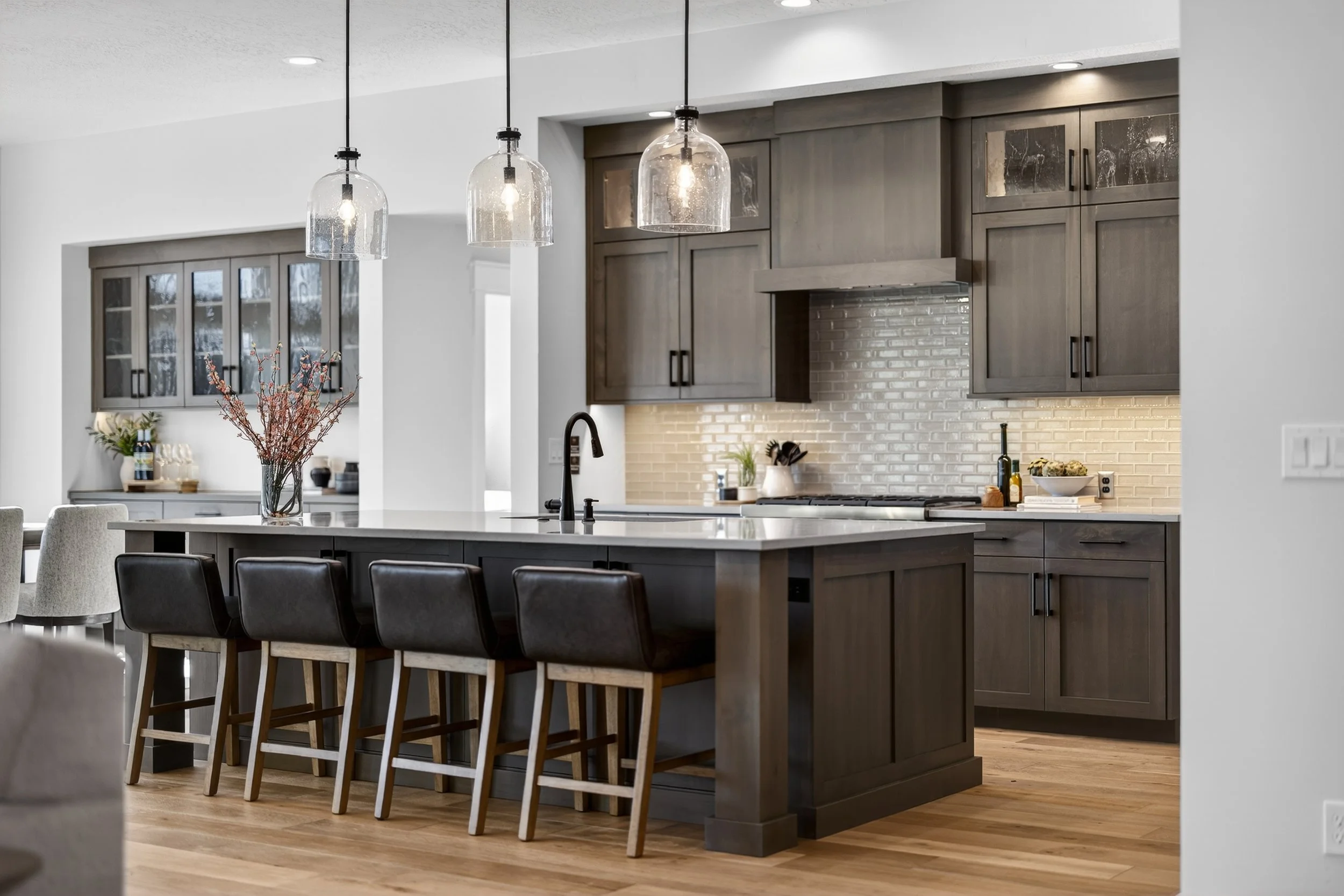Modern kitchen with a central island featuring four barstools with leather seats, gray cabinetry, a white subway tile backsplash, pendant lights hanging above the island, and wooden flooring.