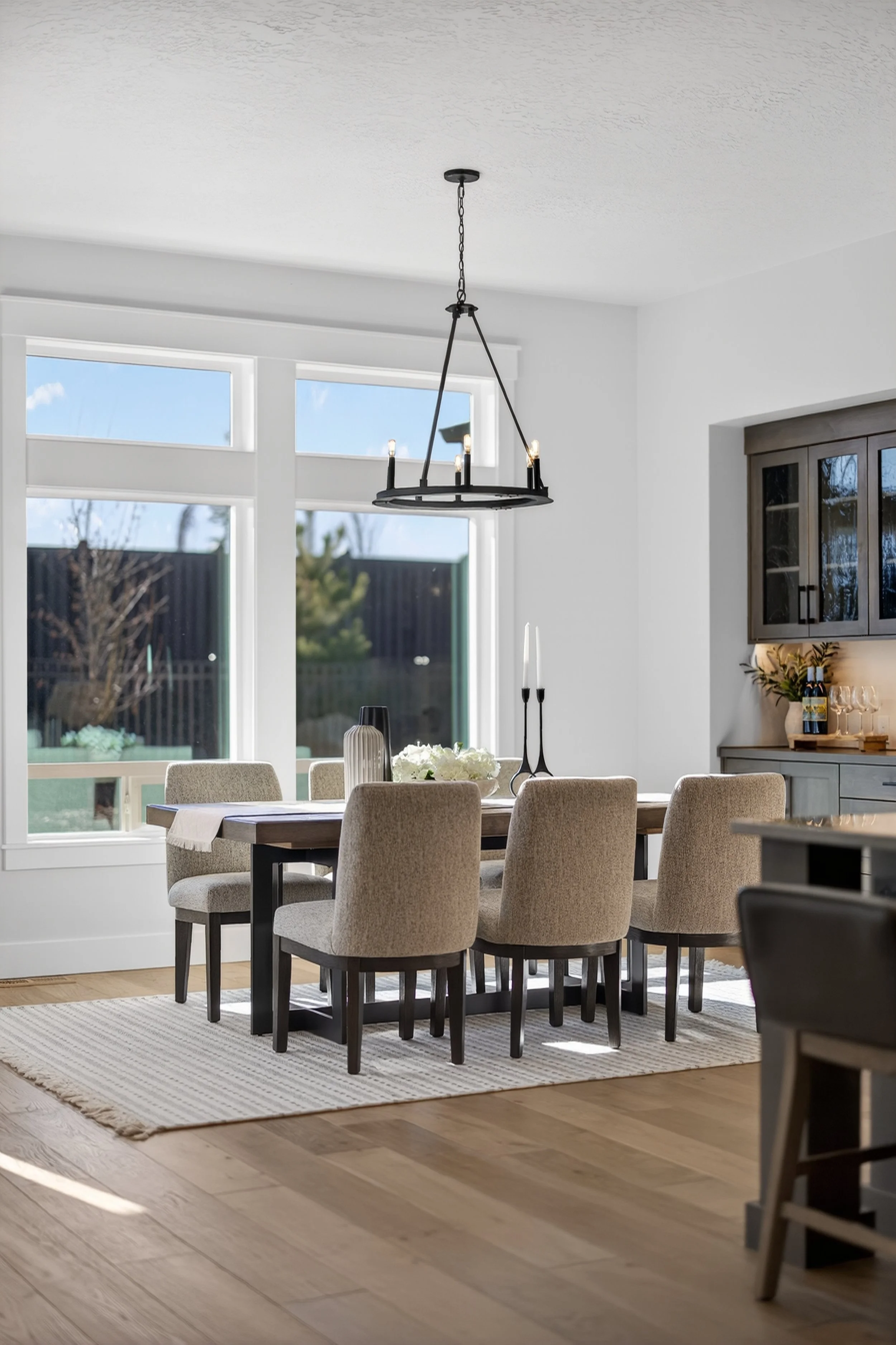 Modern dining room with a wooden table surrounded by beige upholstered chairs, a black metal chandelier hanging above, large window with a garden view, and a white and gray sideboard with decorative items.