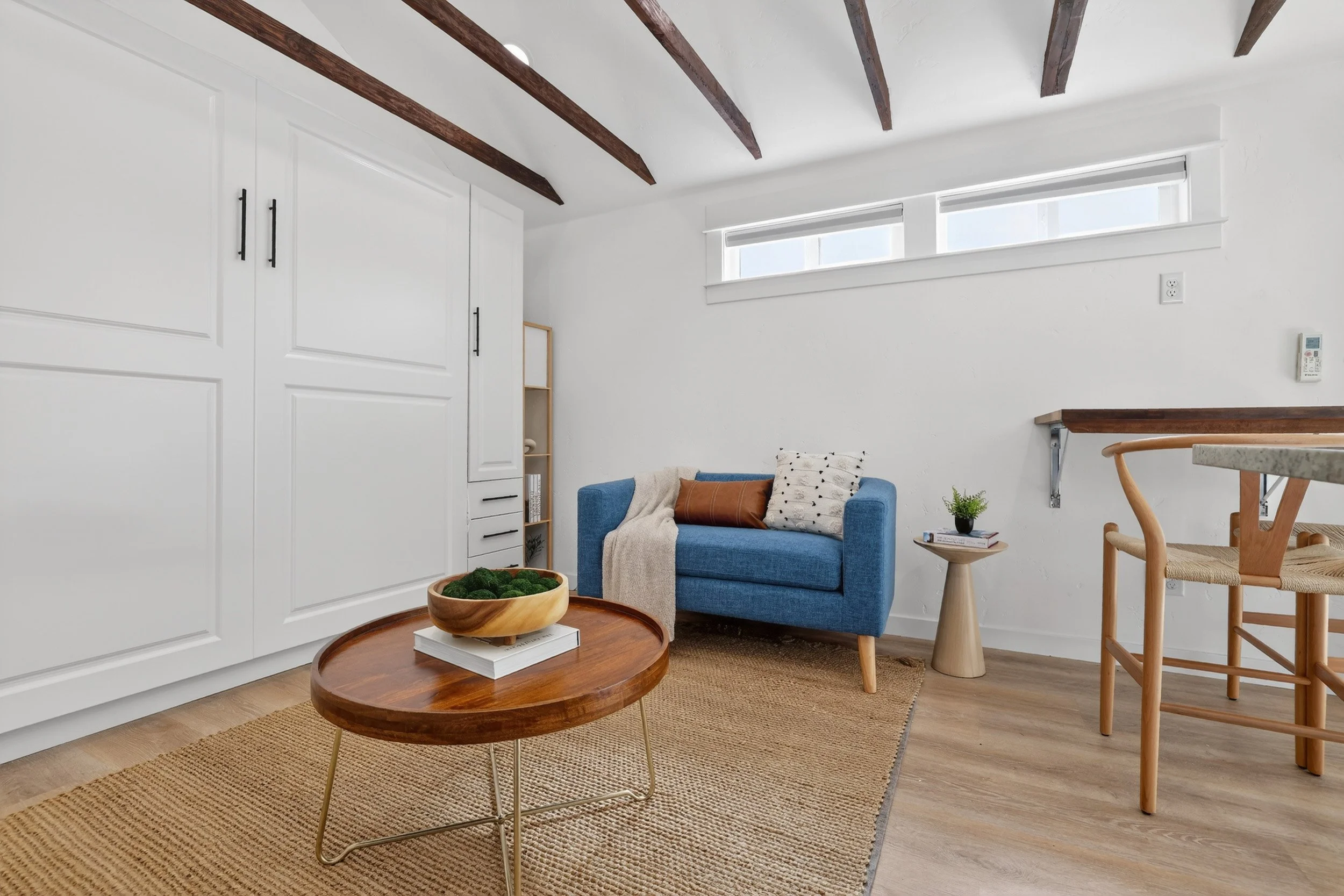 Living room with white walls, wooden ceiling beams, blue sofa with pillows and a throw, wooden coffee table with a bowl and books, small side table with plants and books, and a corner bookshelf.