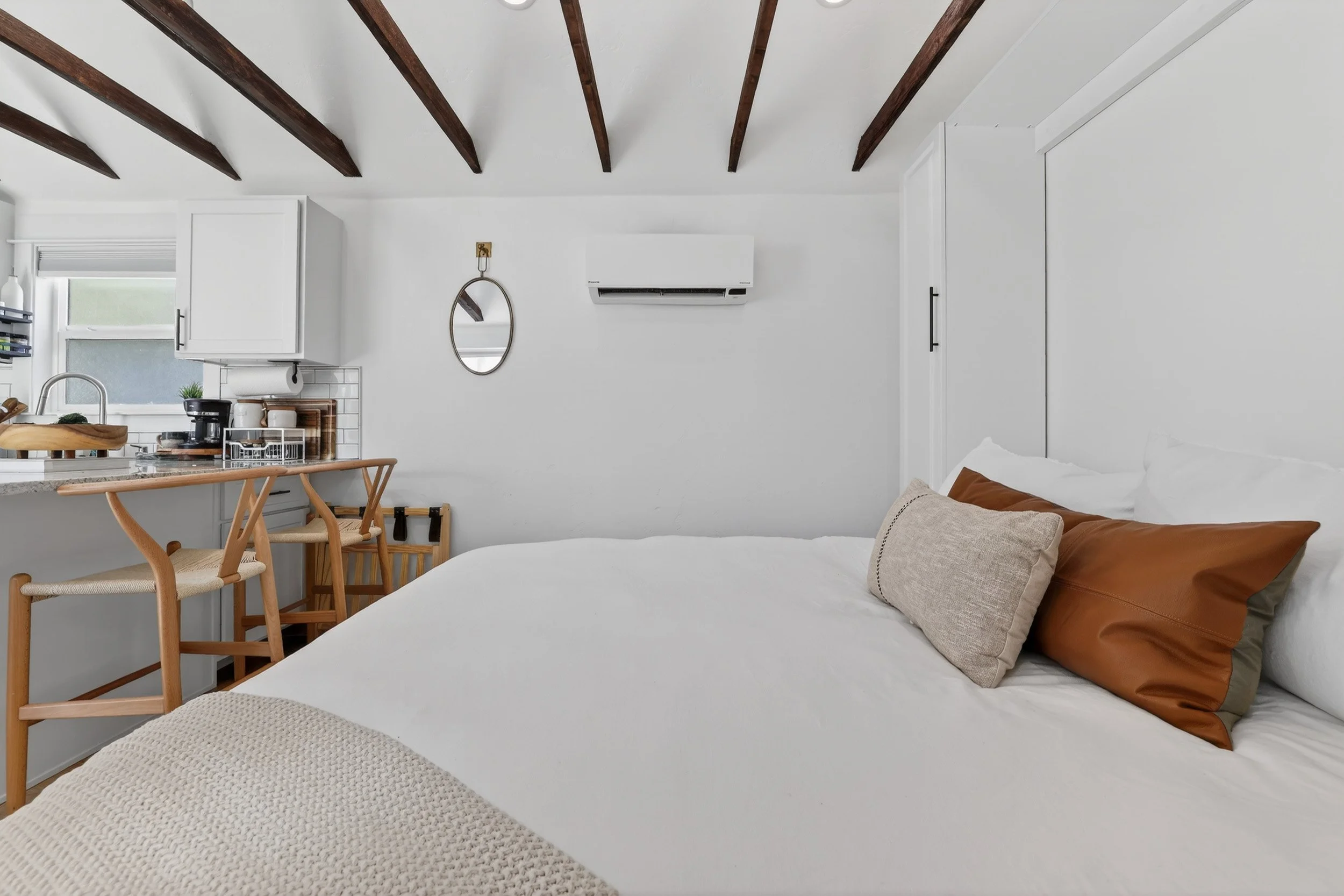 A cozy bedroom with a white bed decorated with two brown pillows and a beige pillow, a small knitted blanket, and a minimalist round mirror on the white wall. There is a window with a small kitchen counter and wooden chairs nearby, and a split air co