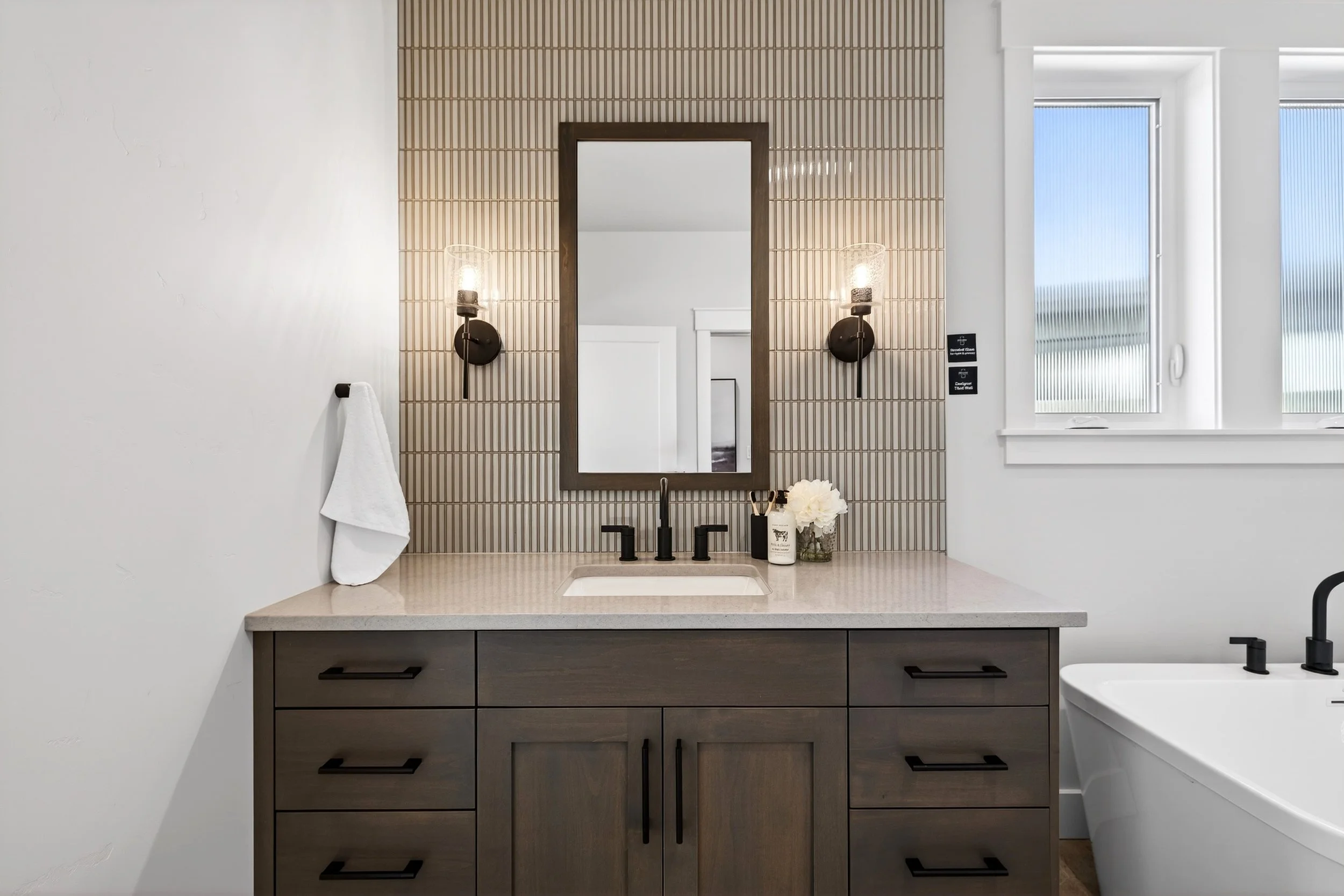 Modern bathroom vanity with a gray wooden cabinet, beige countertop, and black fixtures, with a mirror, wall sconces, and a window.