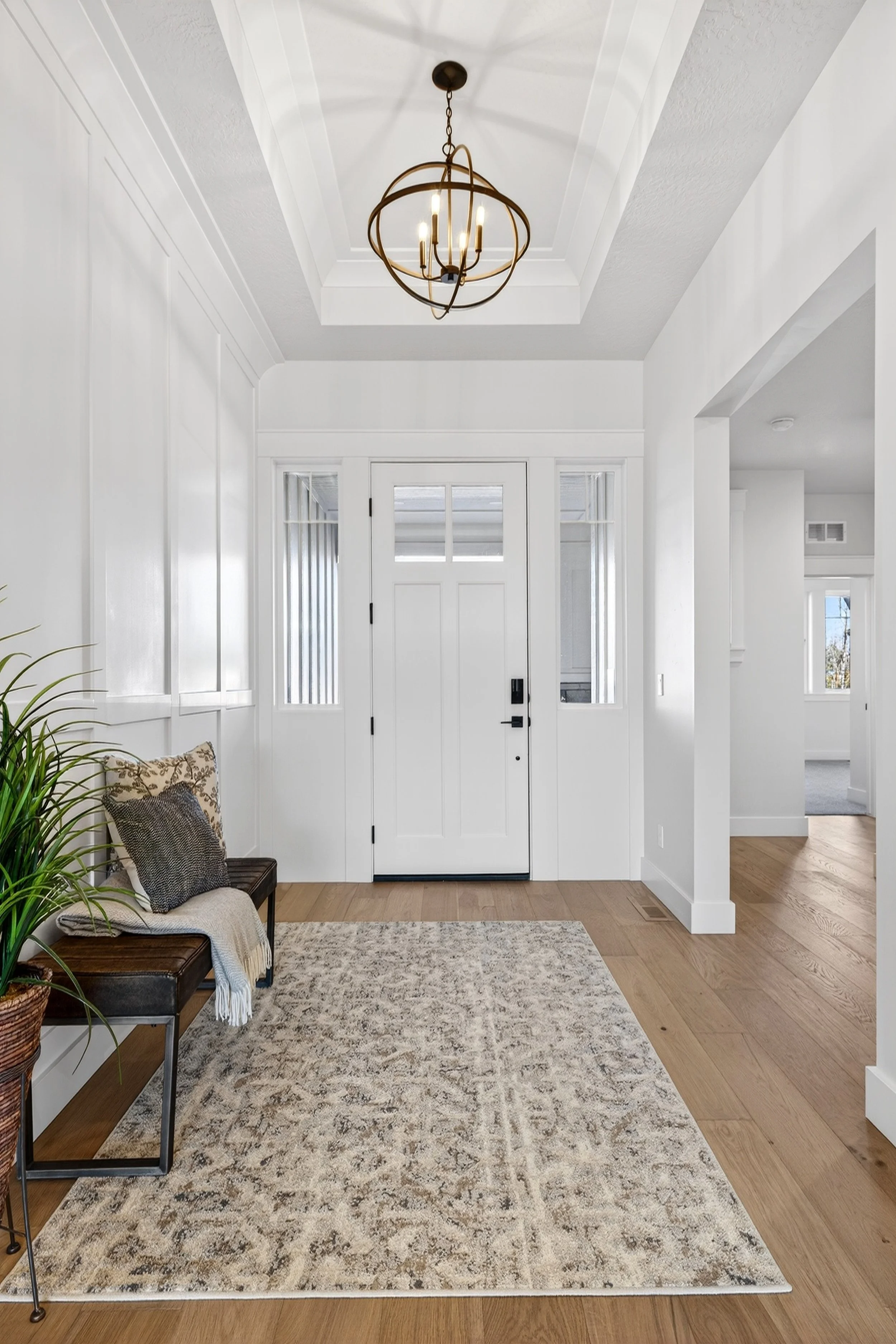 This image shows a bright and inviting foyer with a white front door and two side windows. There is a black bench with patterned pillows and a throw blanket on it, and a large beige patterned rug on the hardwood floor. A modern chandelier hangs from 