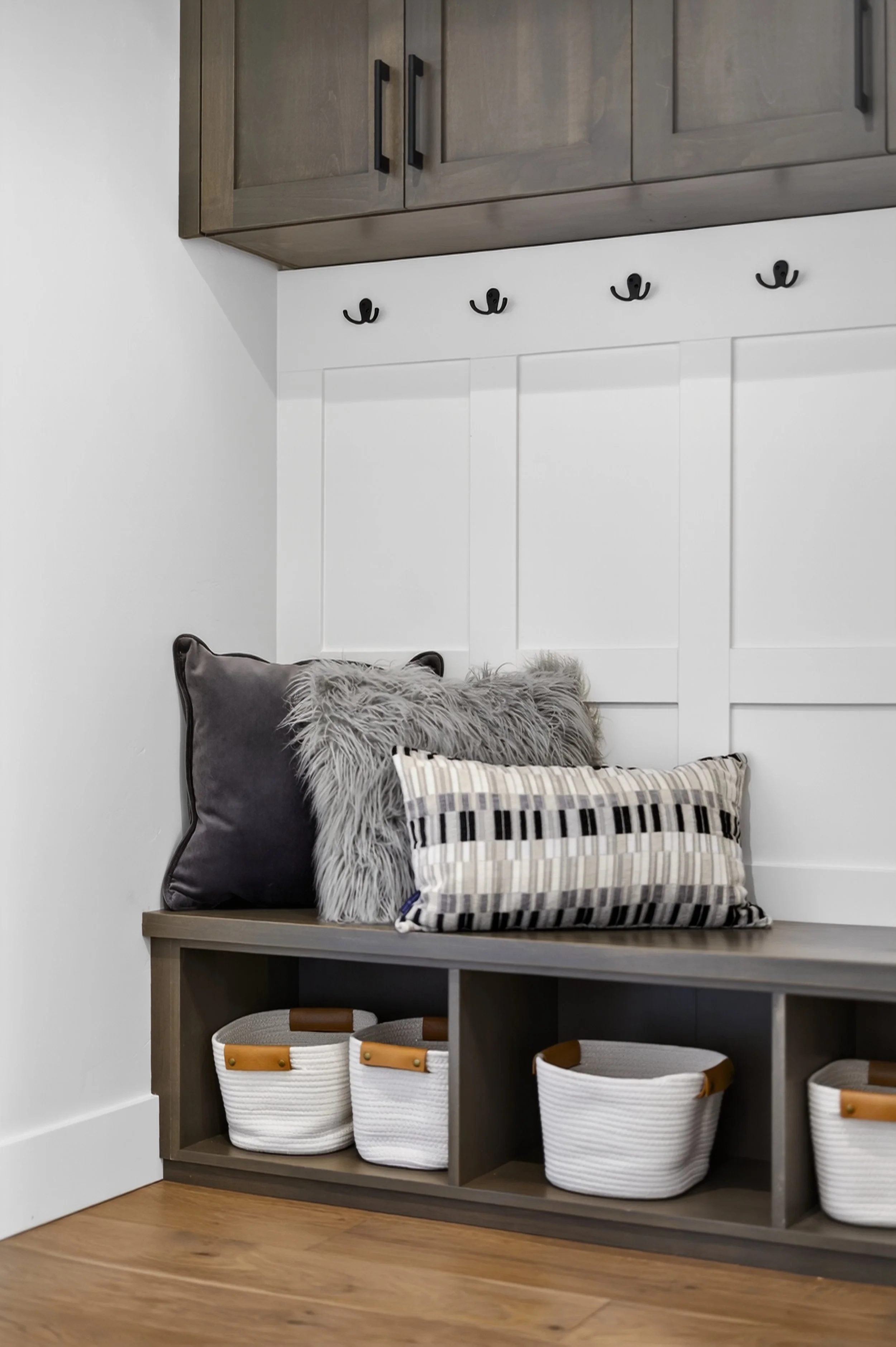 Entryway with a bench, decorative pillows, wall hooks, wooden cabinets, and storage baskets.