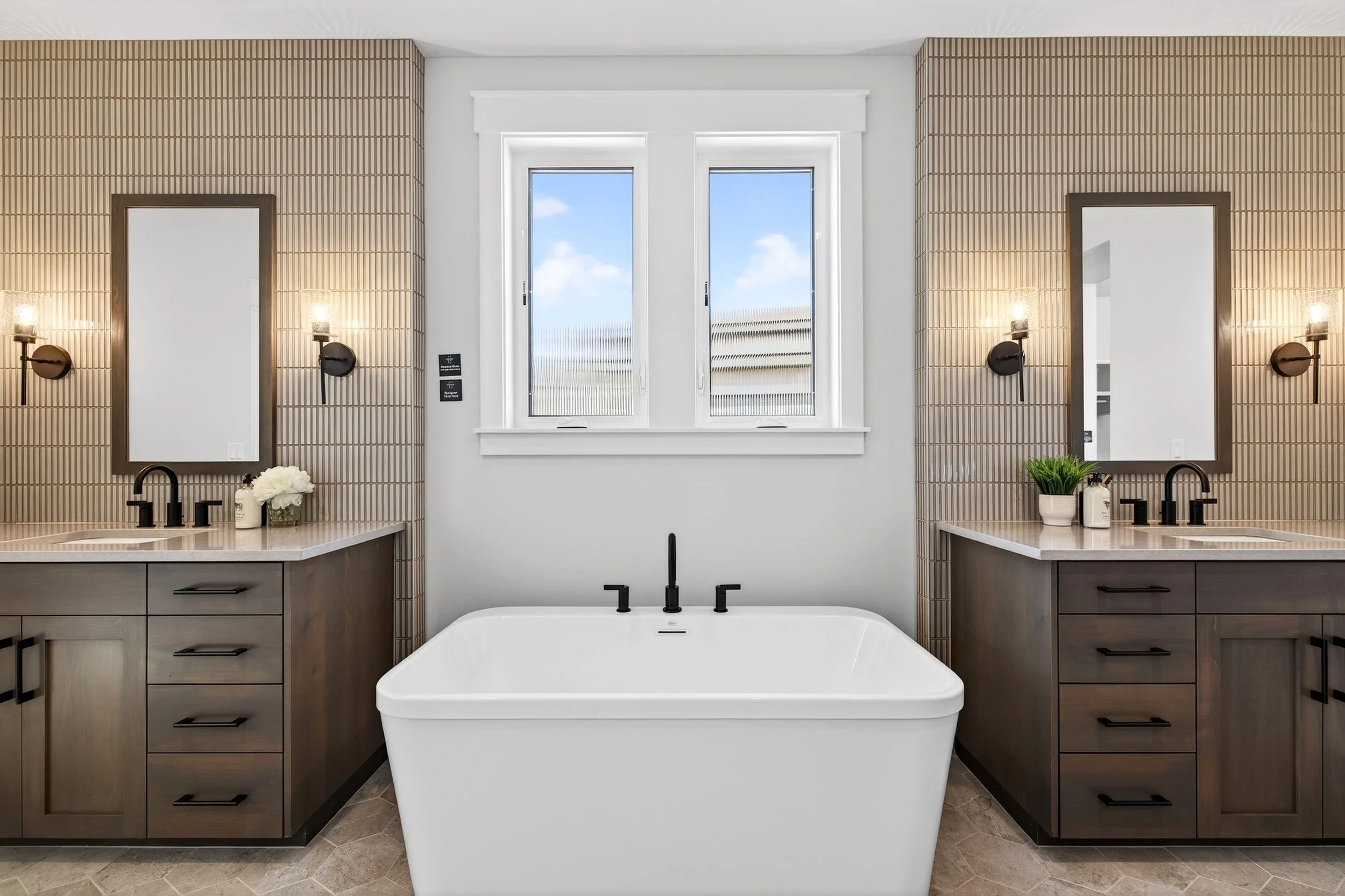 Modern bathroom featuring a freestanding bathtub centered between two vanity stations with sinks, mirrors, and wall-mounted lights, with a window above the bathtub and light-colored tile on the walls and floor.