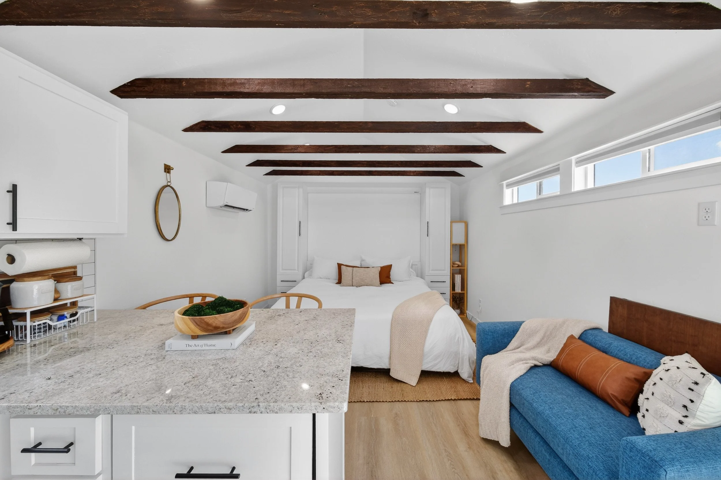 Modern studio apartment with white walls, wooden ceiling beams, a bed with white linens and brown accent pillows, a blue sofa with throw pillows, a granite kitchen island, and a long horizontal window.