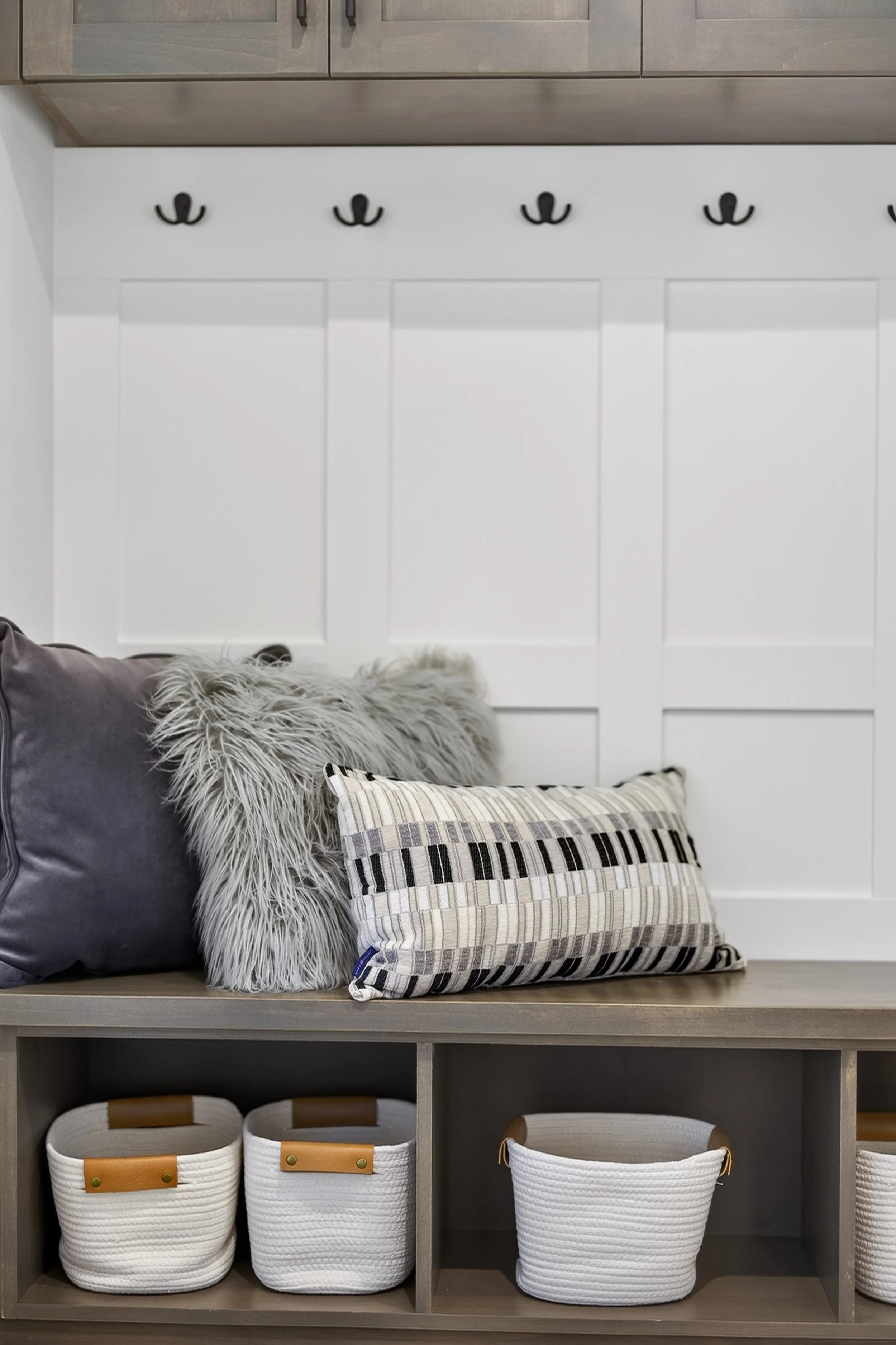 A cozy mudroom bench with decorative pillows and storage baskets below. The bench is against a white paneled wall with black coat hooks above.