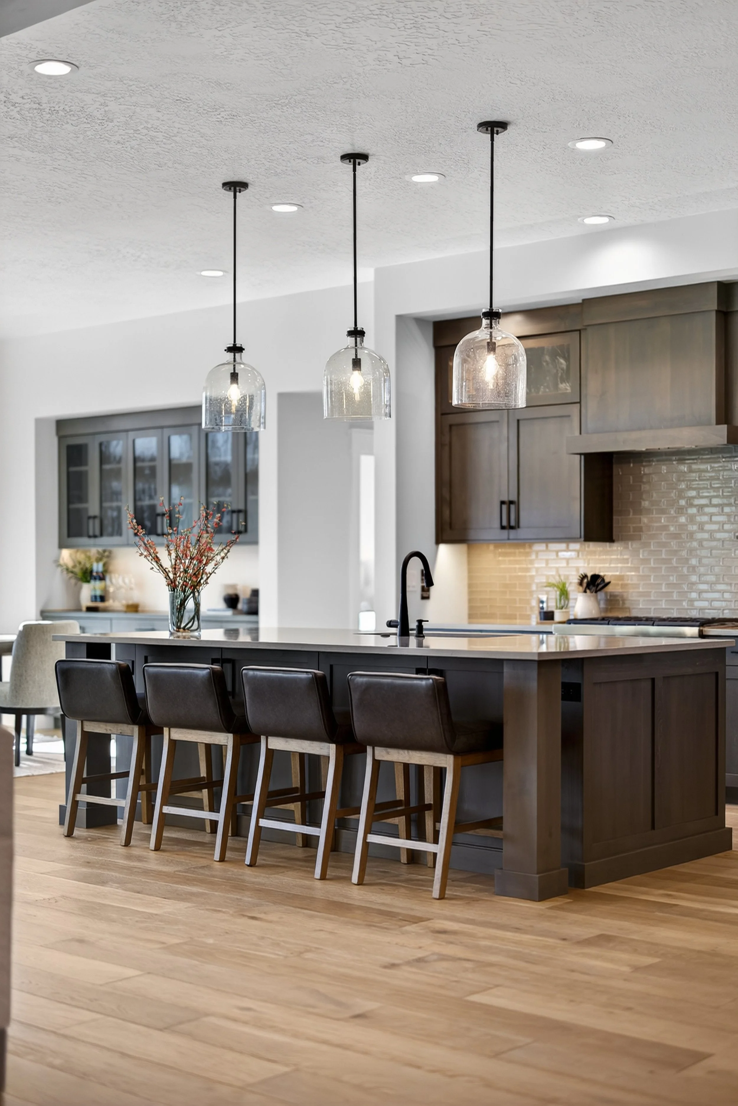 Modern kitchen with a large island, black chairs, pendant lighting, wooden cabinets, and hardwood floors.