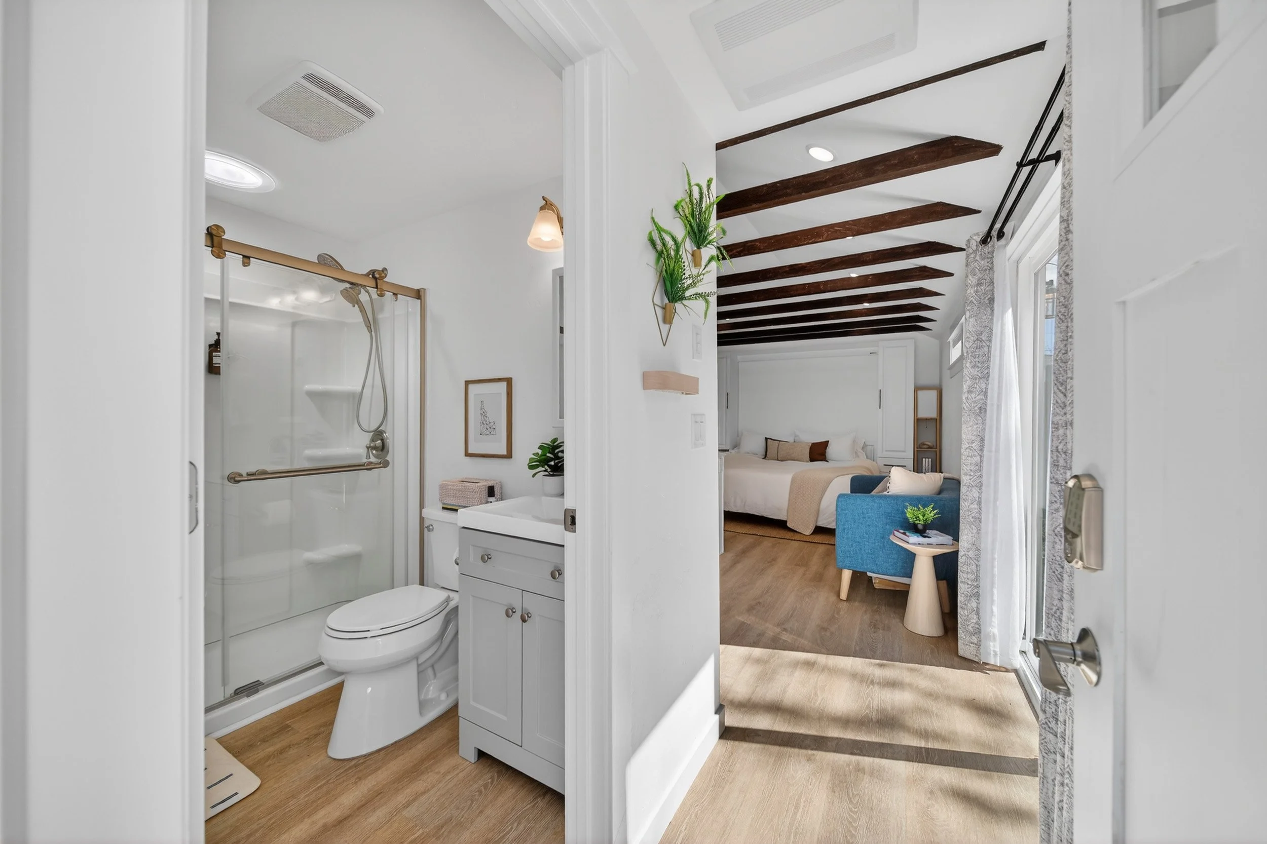 Bathroom with a toilet and a shower, view towards the bedroom with a bed, blue armchair, side table, and decorative plants.